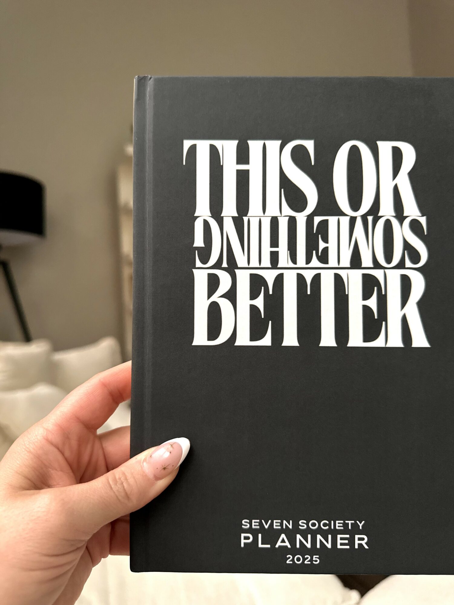 Hand holding a black planner with white text reading 'This or that, better, seven society planner 2025'.