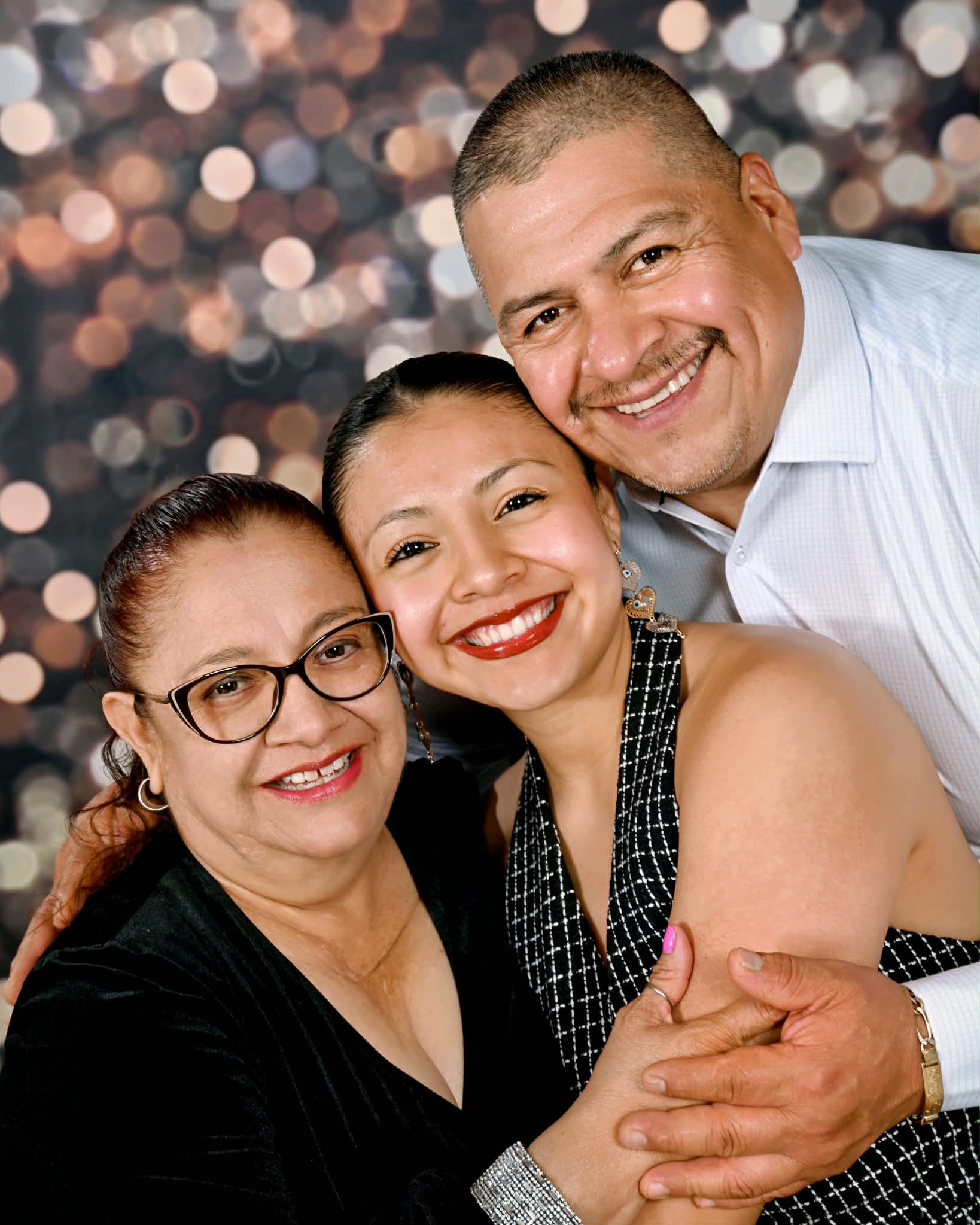 Three smiling people, two women and one man, close together, with a blurred festive background.