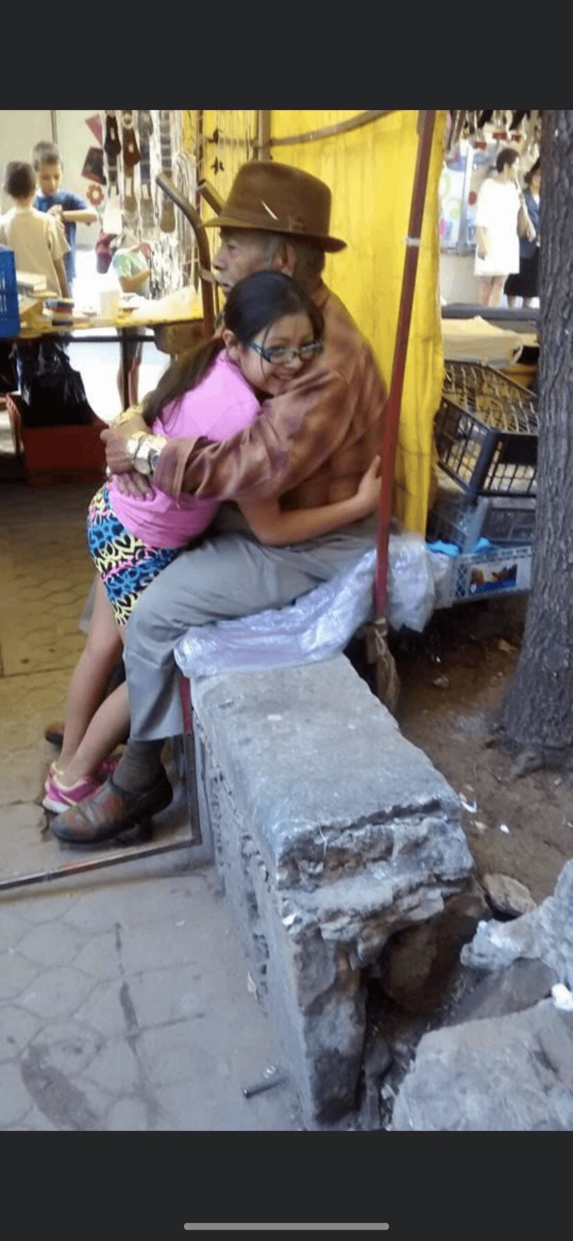 Two women hugging, one sitting on a concrete block, in a market with yellow curtains and people in the background.