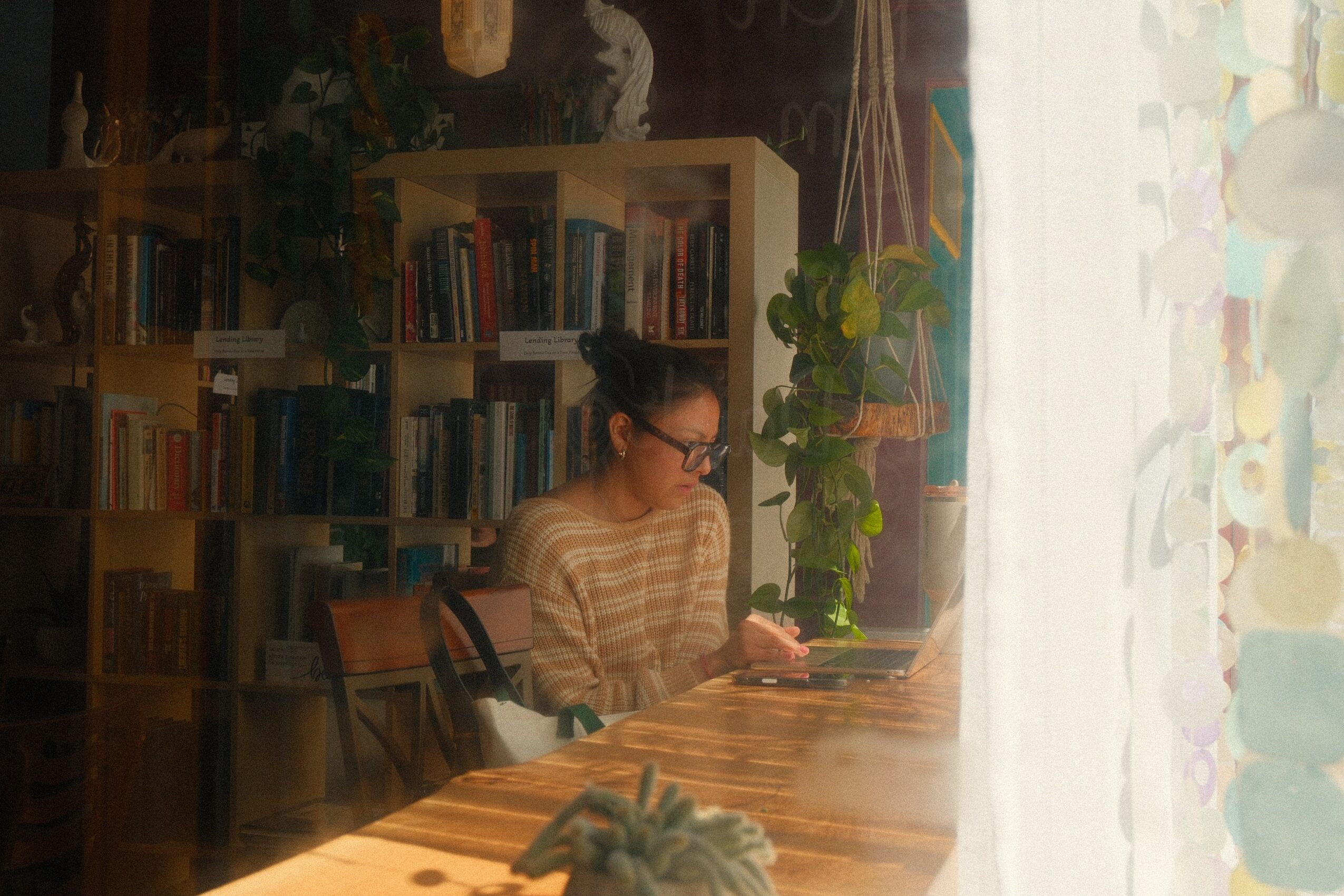 A woman with glasses and a striped shirt sits at a wooden table reading a book in a room with bookshelves and plants.