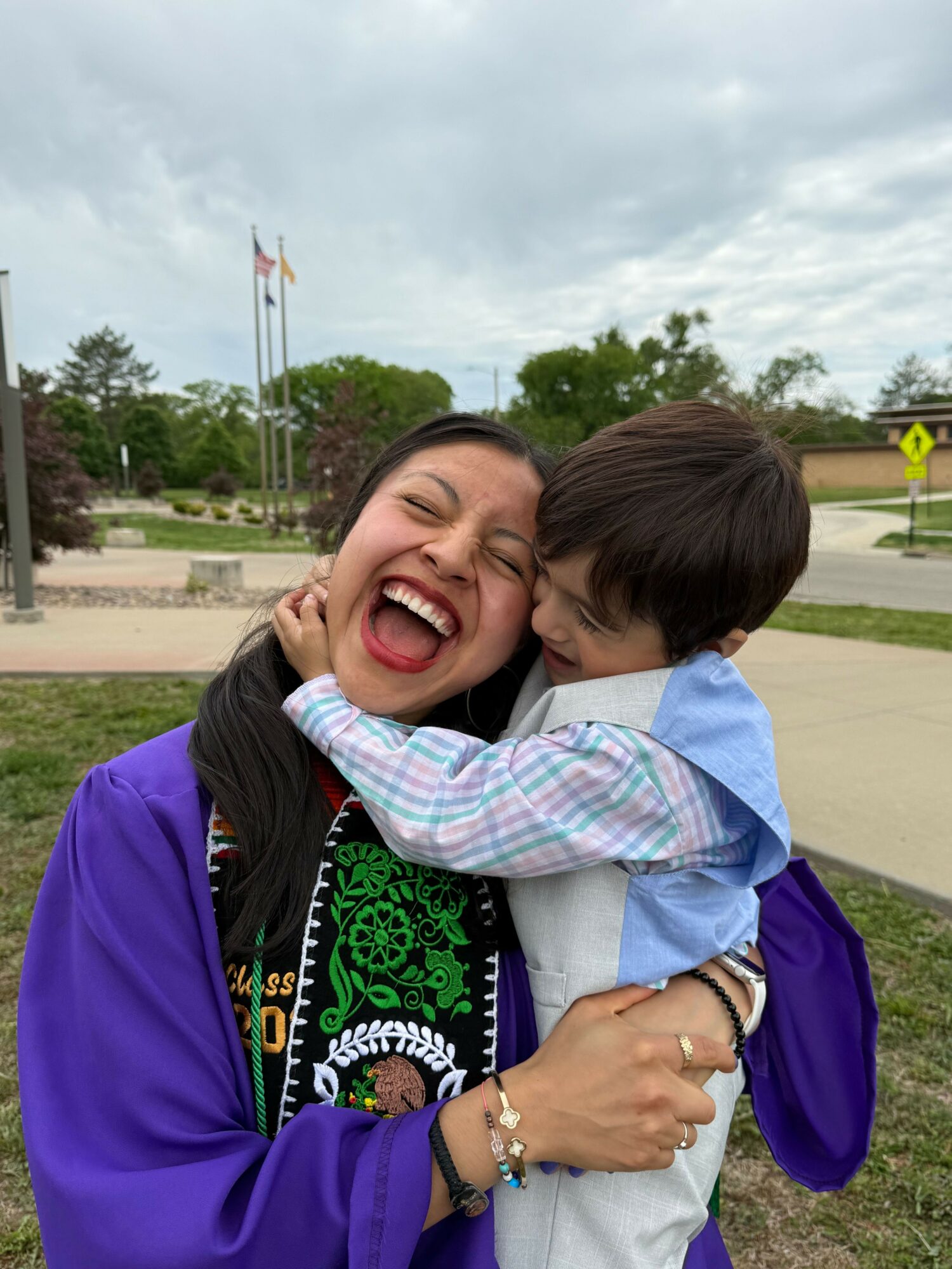 A woman and a young boy are smiling and hugging outdoors on a cloudy day.