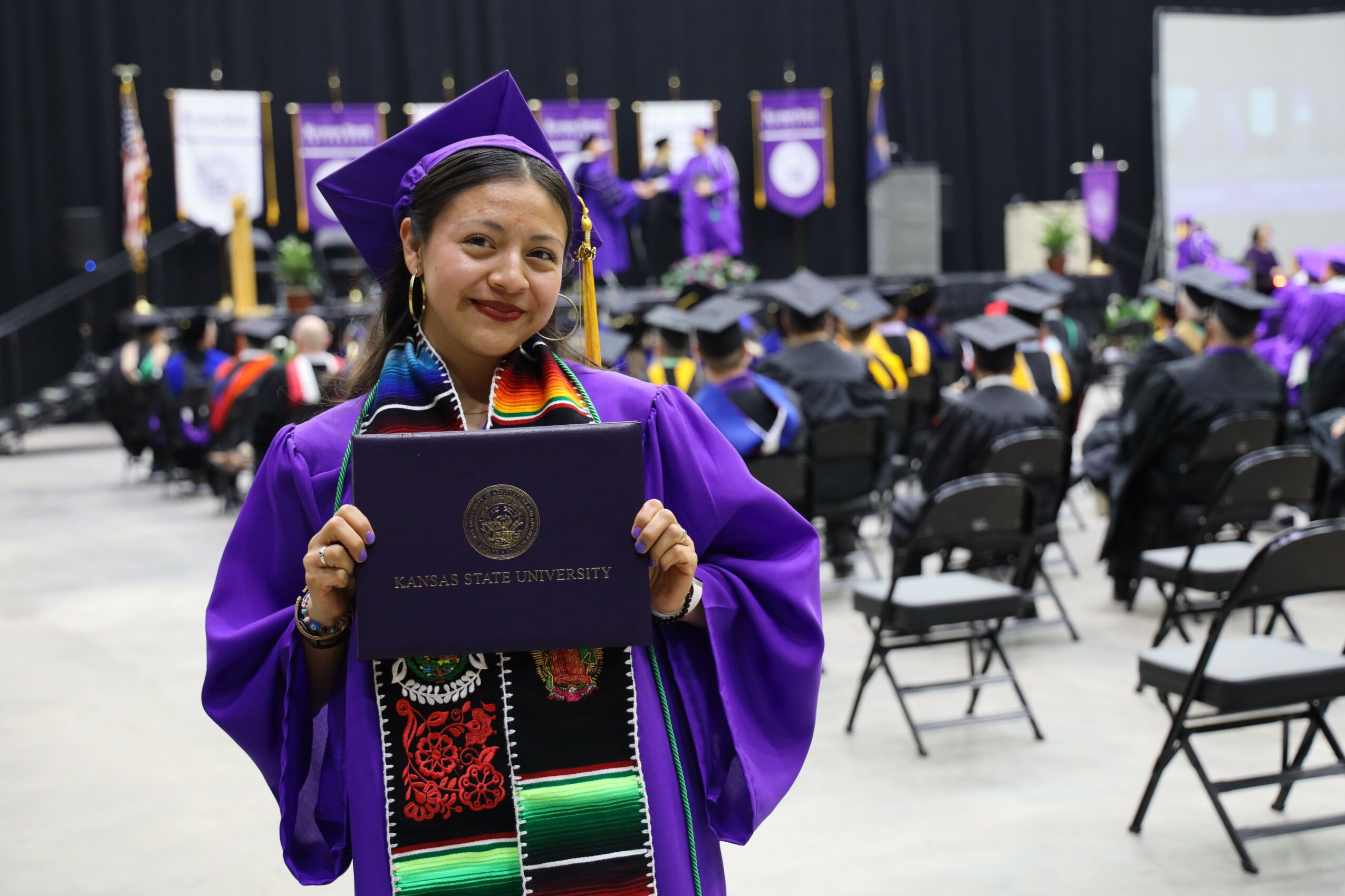 Young woman in graduation gown and cap holding diploma, smiling at ceremony with seated audience and stage in background.