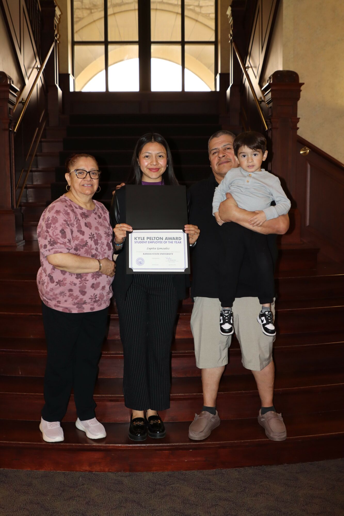 Three women standing indoors, one holding a certificate, with wooden floors and large windows in the background.