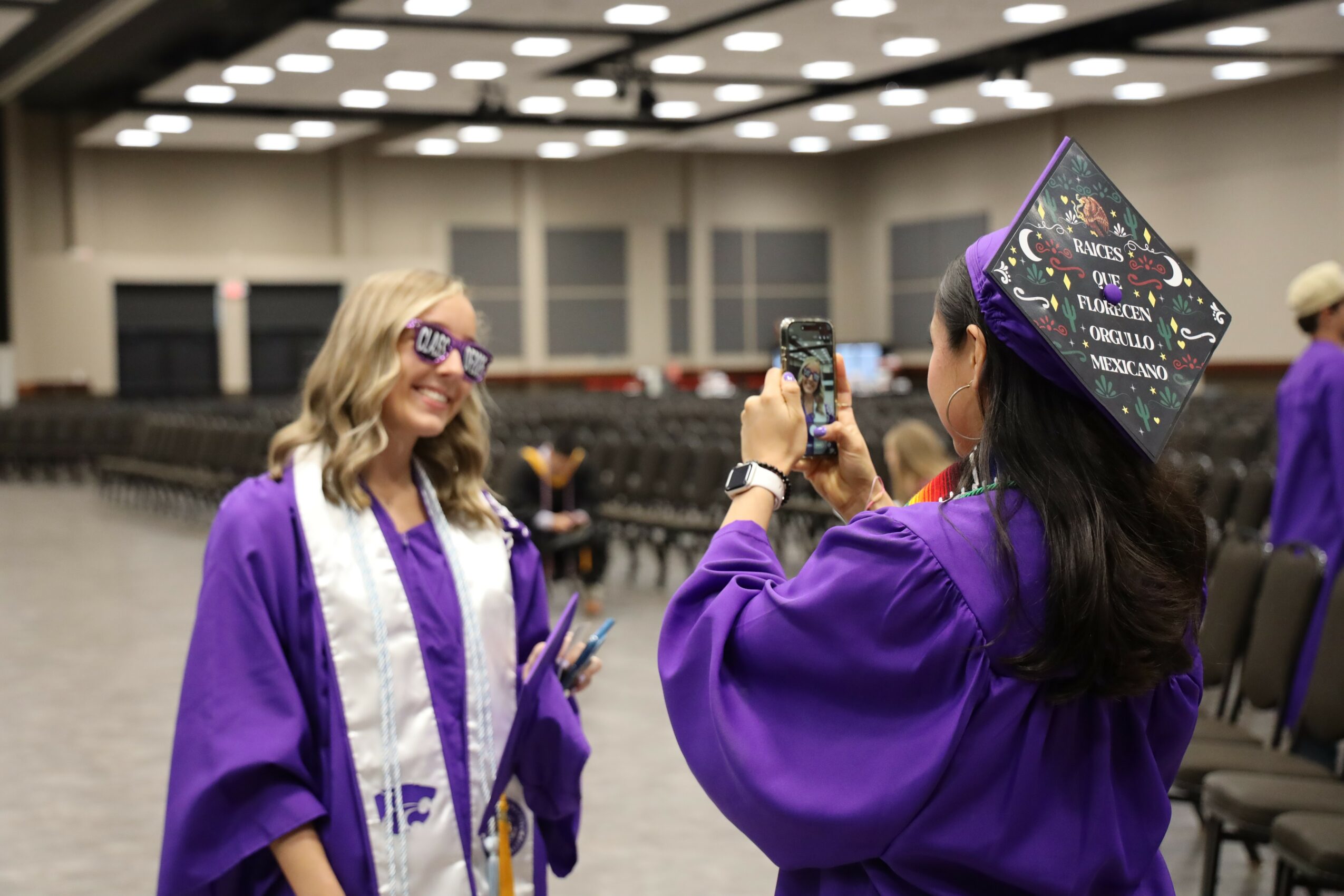 Two women in purple graduation gowns and caps, one taking a photo of the other, in an indoor ceremony hall.