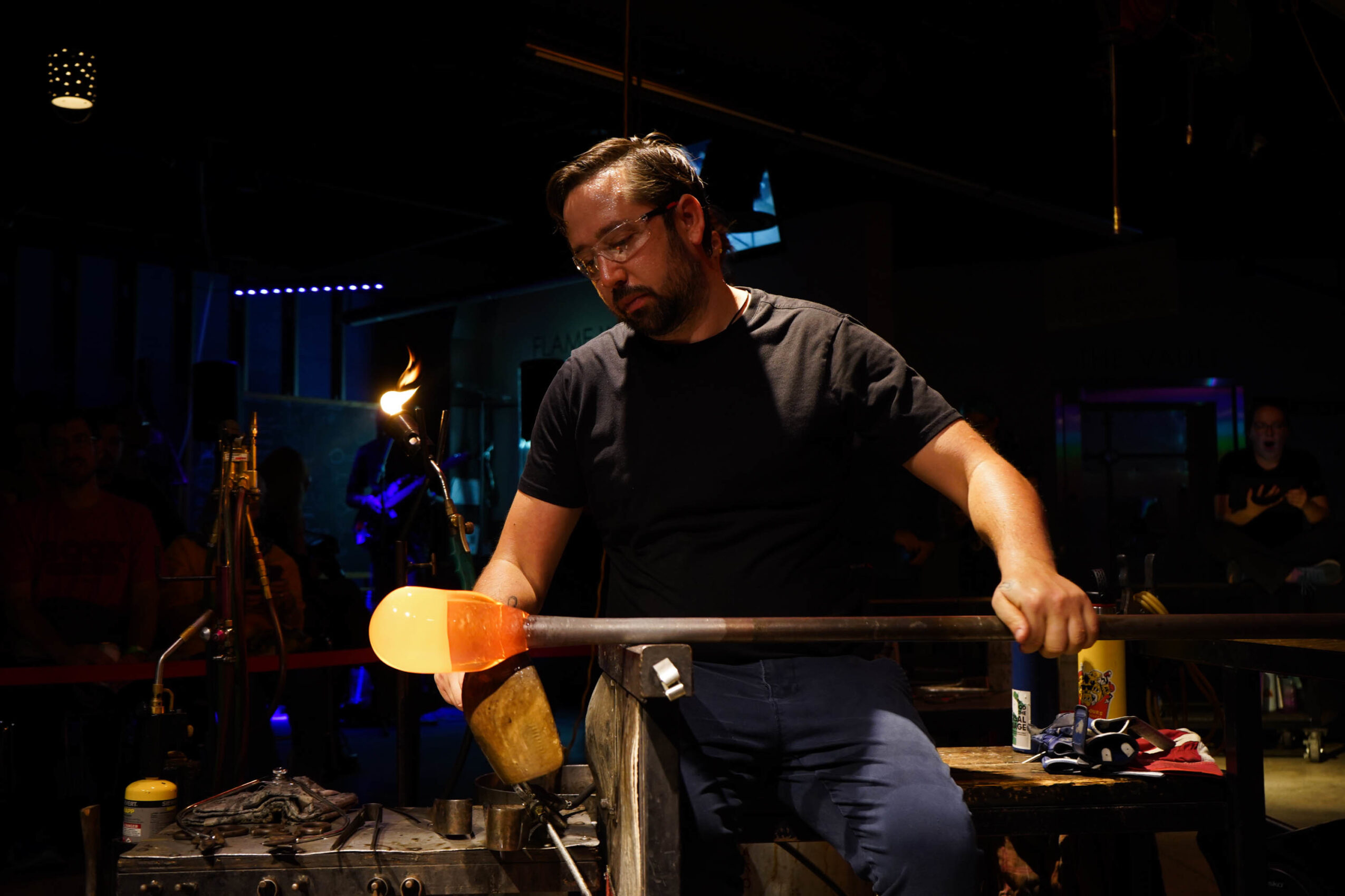 Person with glasses and beard holding a long stick in a dimly lit room with equipment and people in the background.