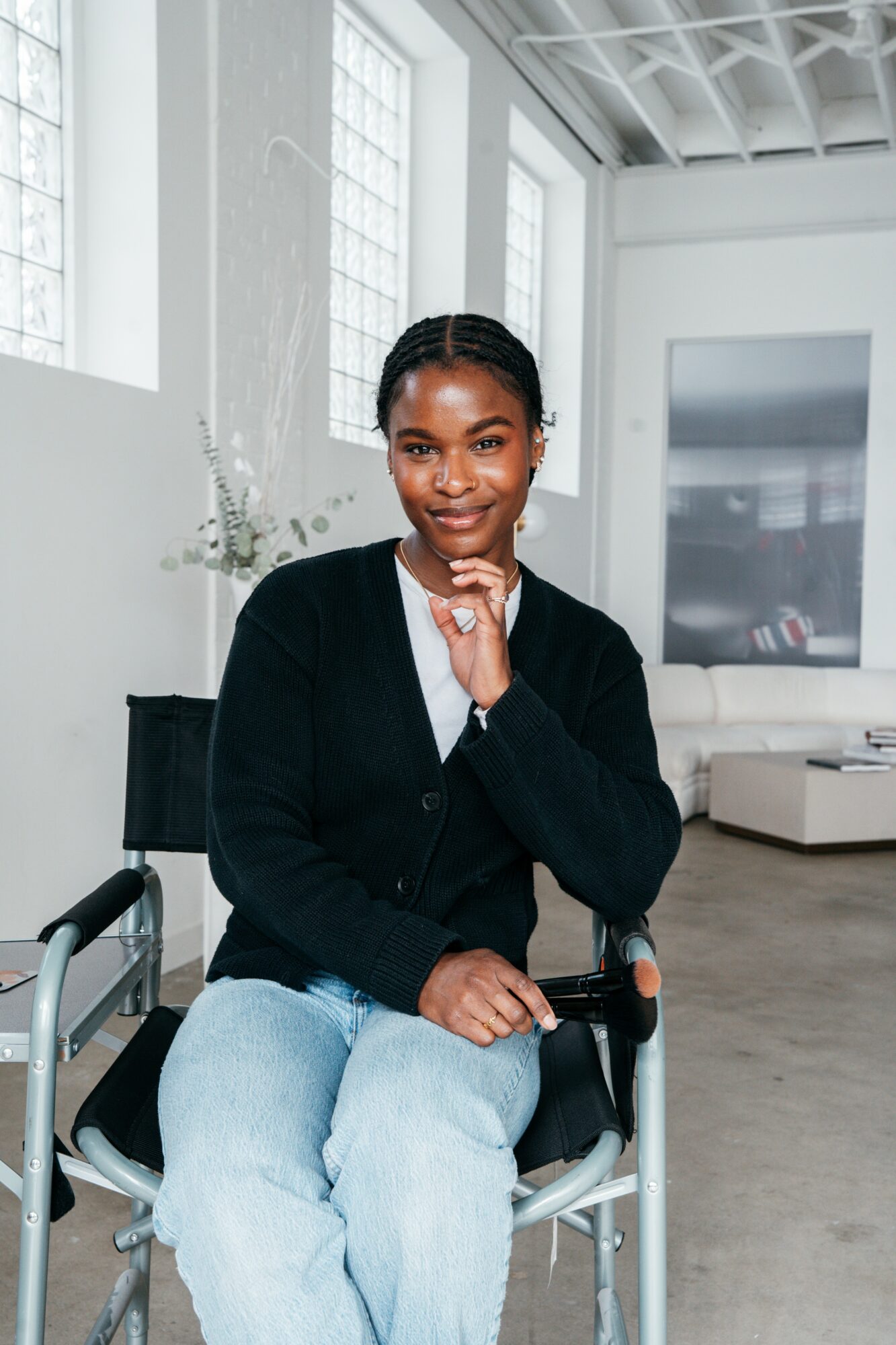Woman sitting in a modern, bright room with large windows, wearing a black cardigan and light jeans, smiling.