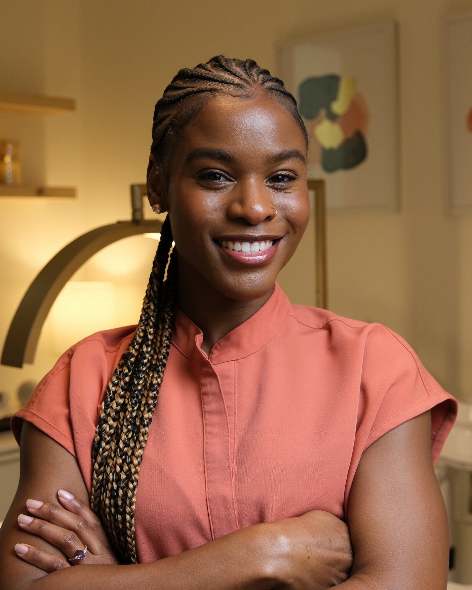 Smiling woman with braided hair wearing a coral-colored top, arms crossed, in a warmly lit room with artwork on the wall.