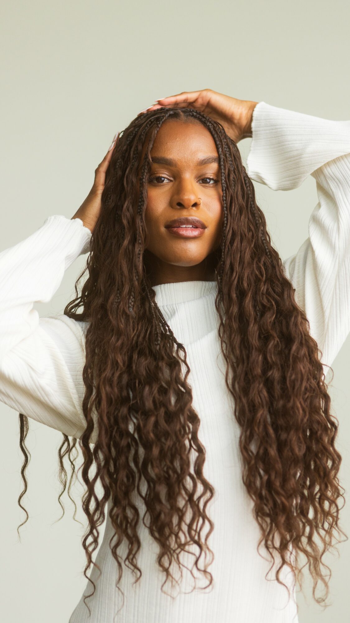 Woman with long, curly hair wearing a white sweater, posing with hands on her head, smiling slightly.
