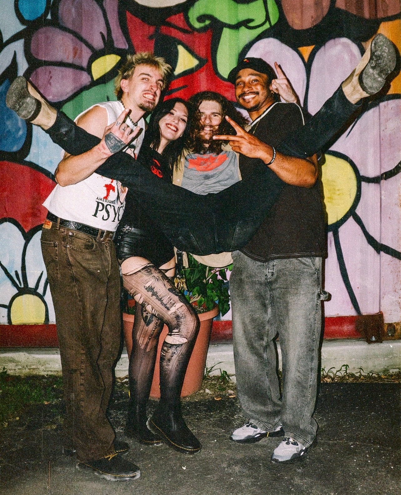 Four people posing in front of a colorful mural, with one person being lifted horizontally by the others, all smiling.