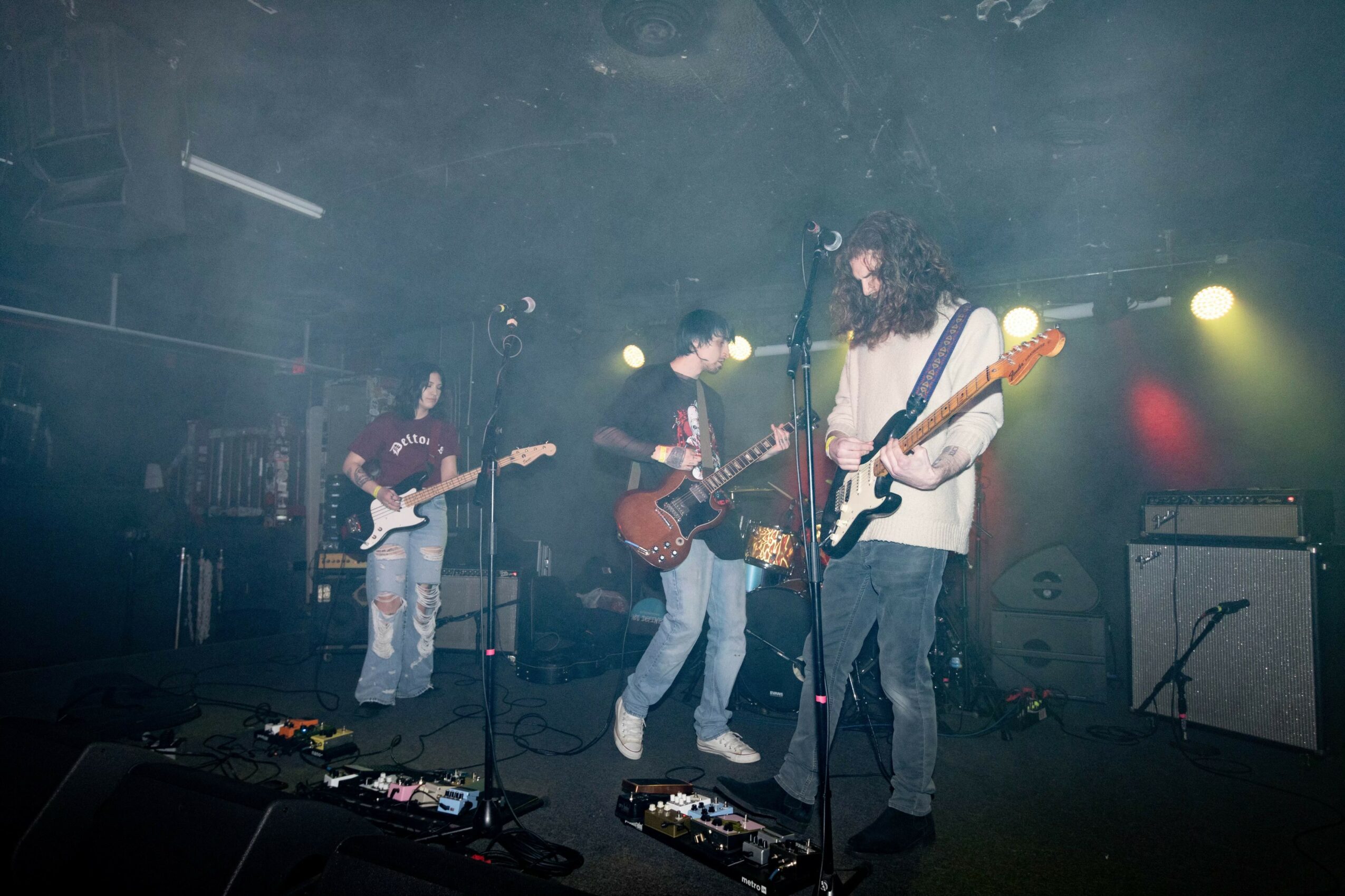 Four musicians perform on stage with guitars, microphones, and equipment, illuminated by stage lights in a dark venue.