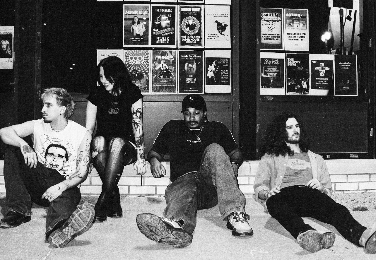 Four people sitting and standing outside a building with posters on the window, at night.