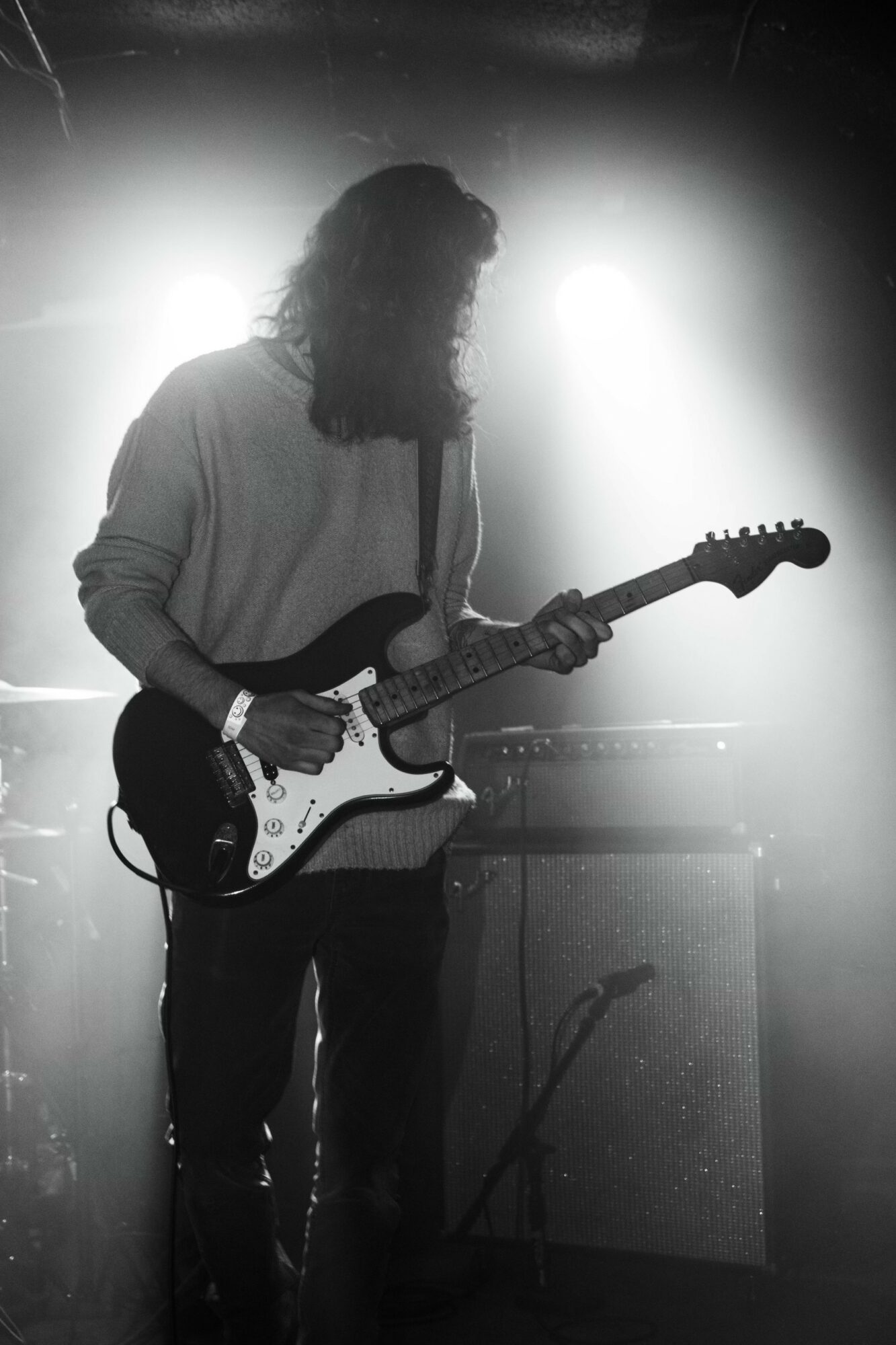 Person playing electric guitar on stage with bright backlight, long hair, and casual clothing.