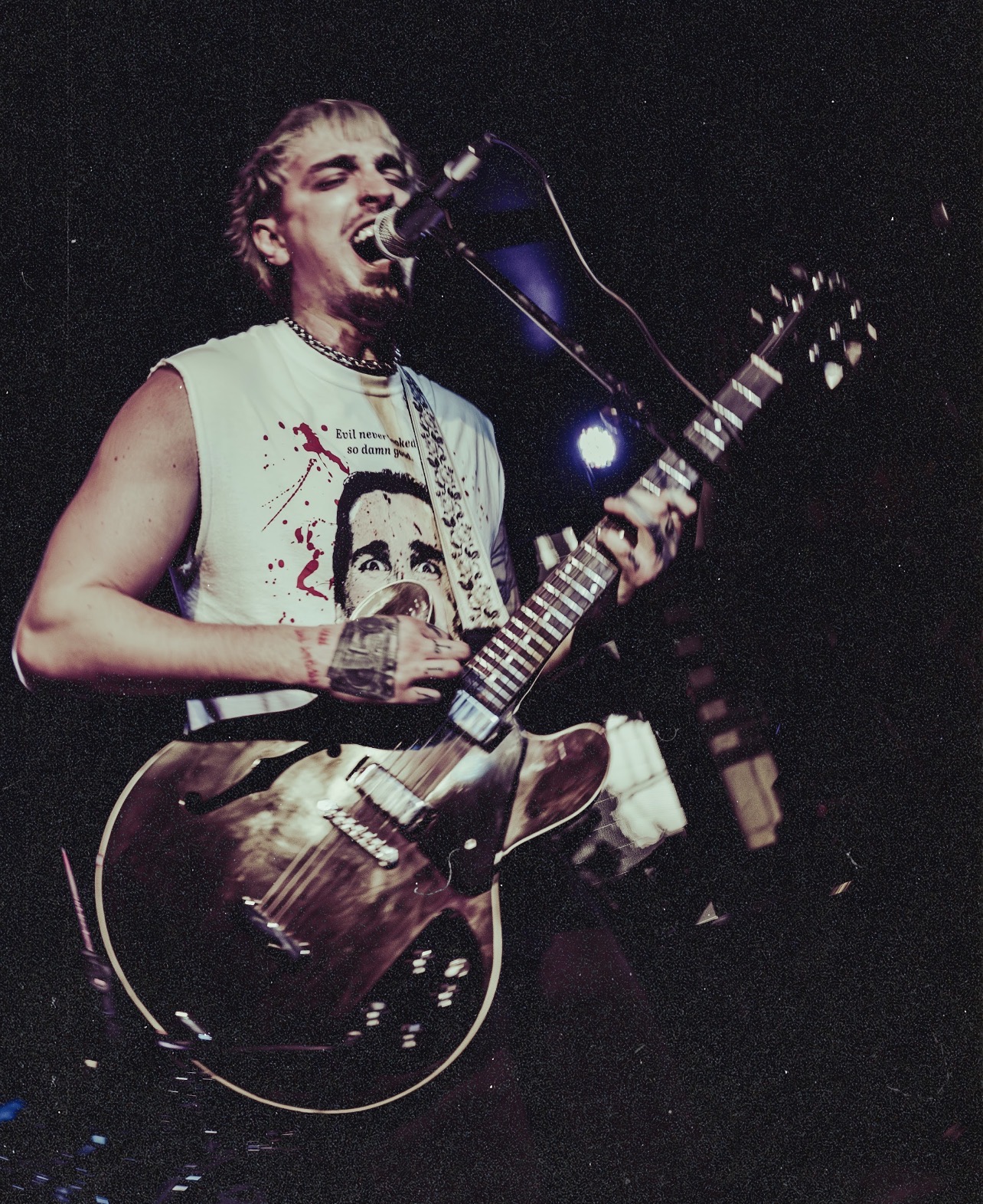 Person singing into a microphone while playing an electric guitar on stage, wearing a sleeveless shirt with a graphic design.