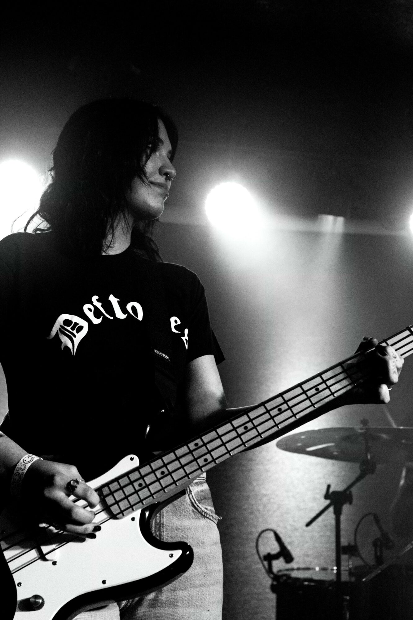 Person playing bass guitar on stage with bright lights behind, dark background, long hair, wearing a black shirt.