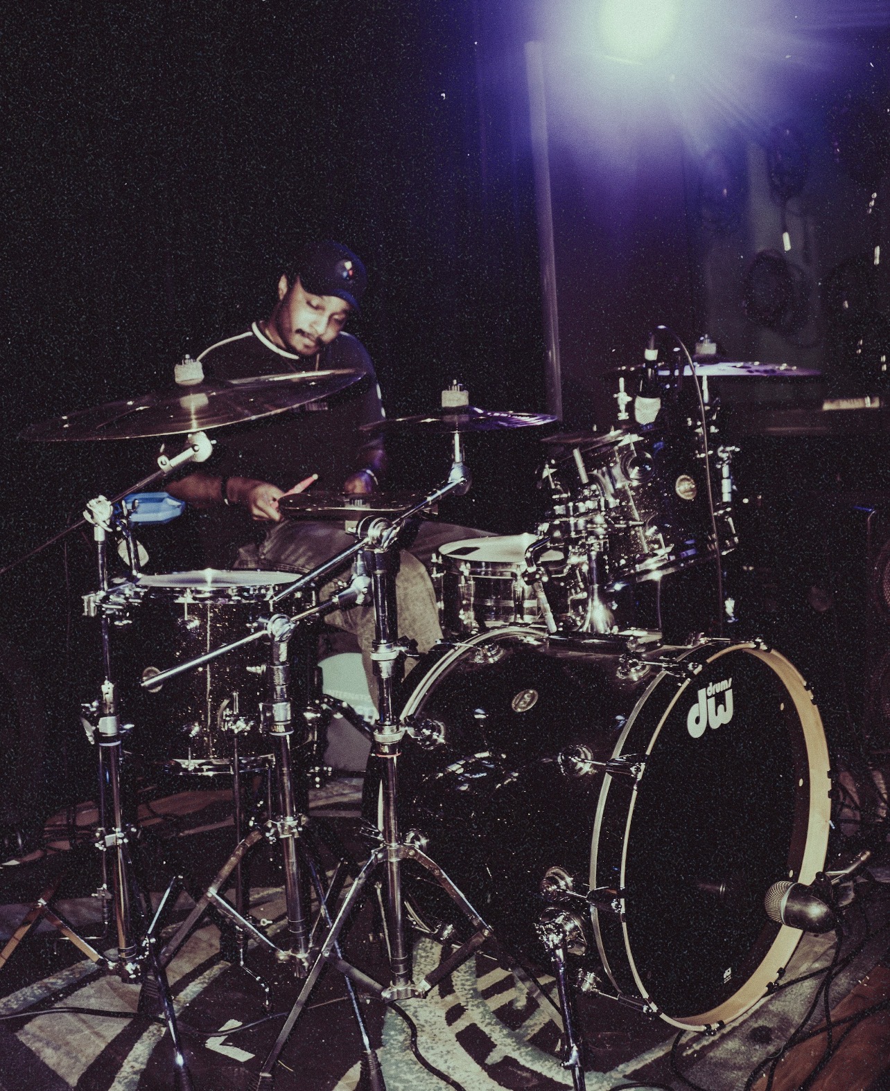 A person playing a drum set on stage with a bright light overhead, dark background, and musical equipment visible.