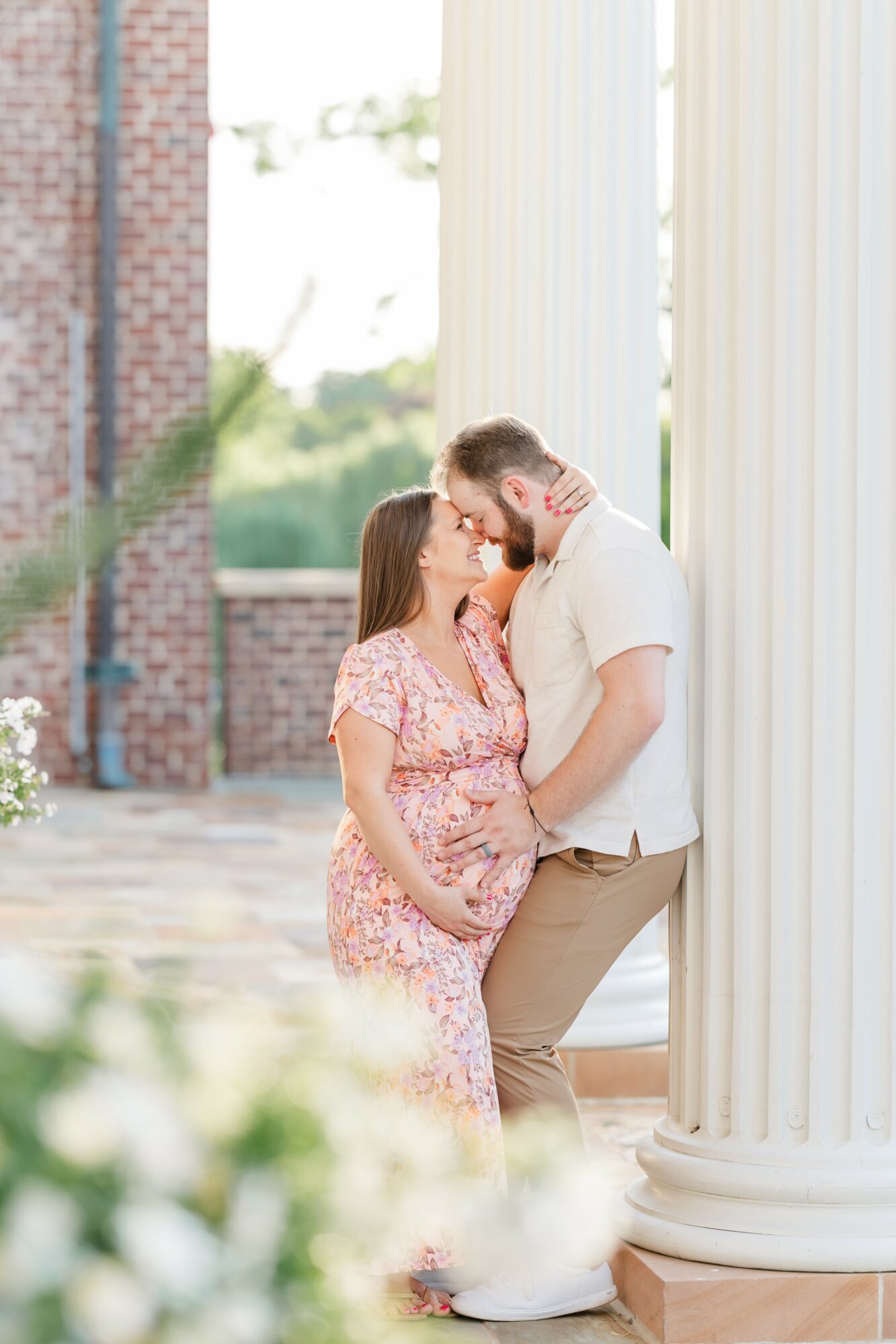 Pregnant woman and man share a kiss near white columns outdoors, with greenery and brick wall in background.