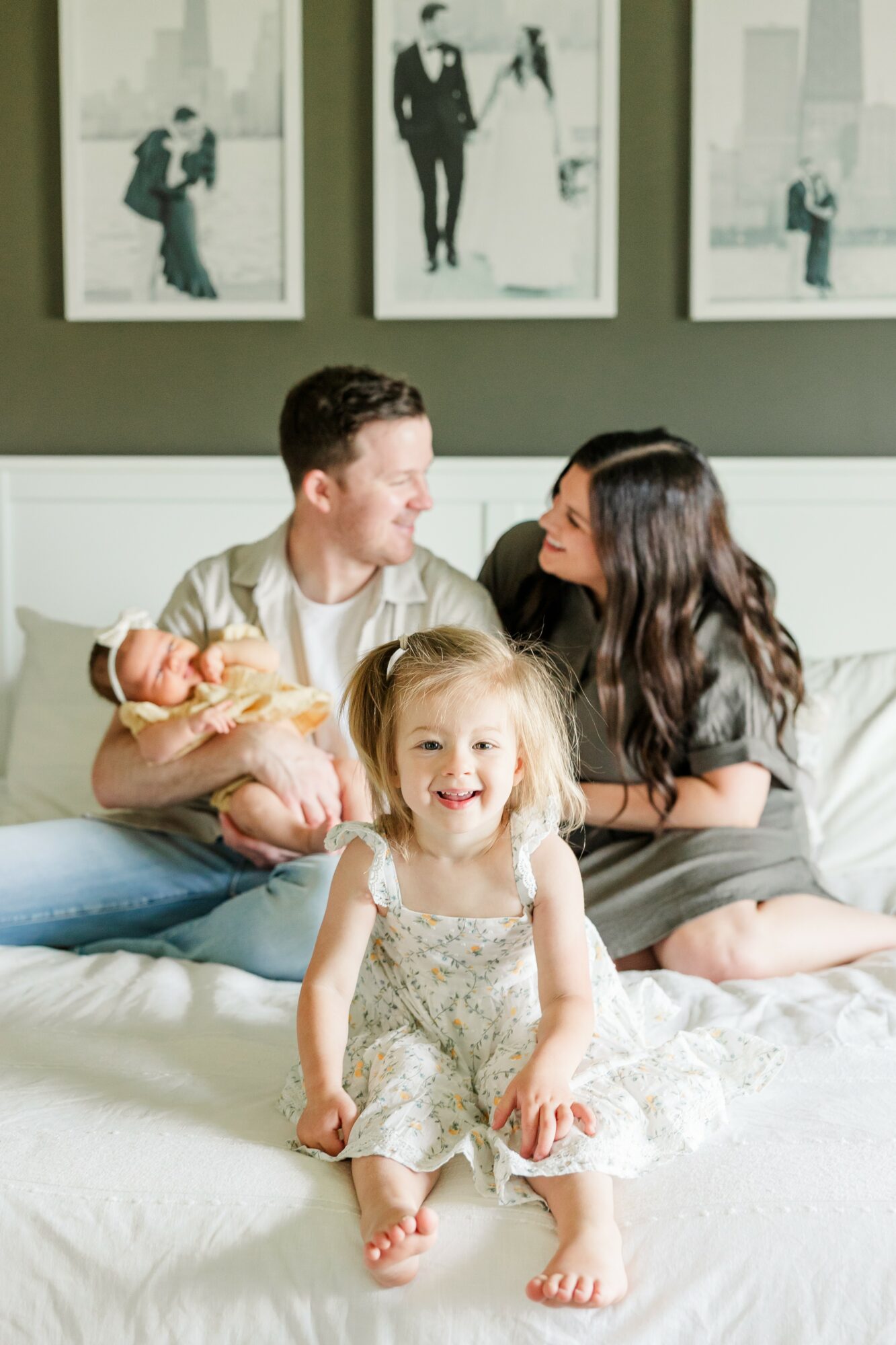 A family of three sitting on a bed with a young girl in front, smiling at the camera, in a room with framed photos on the wall.