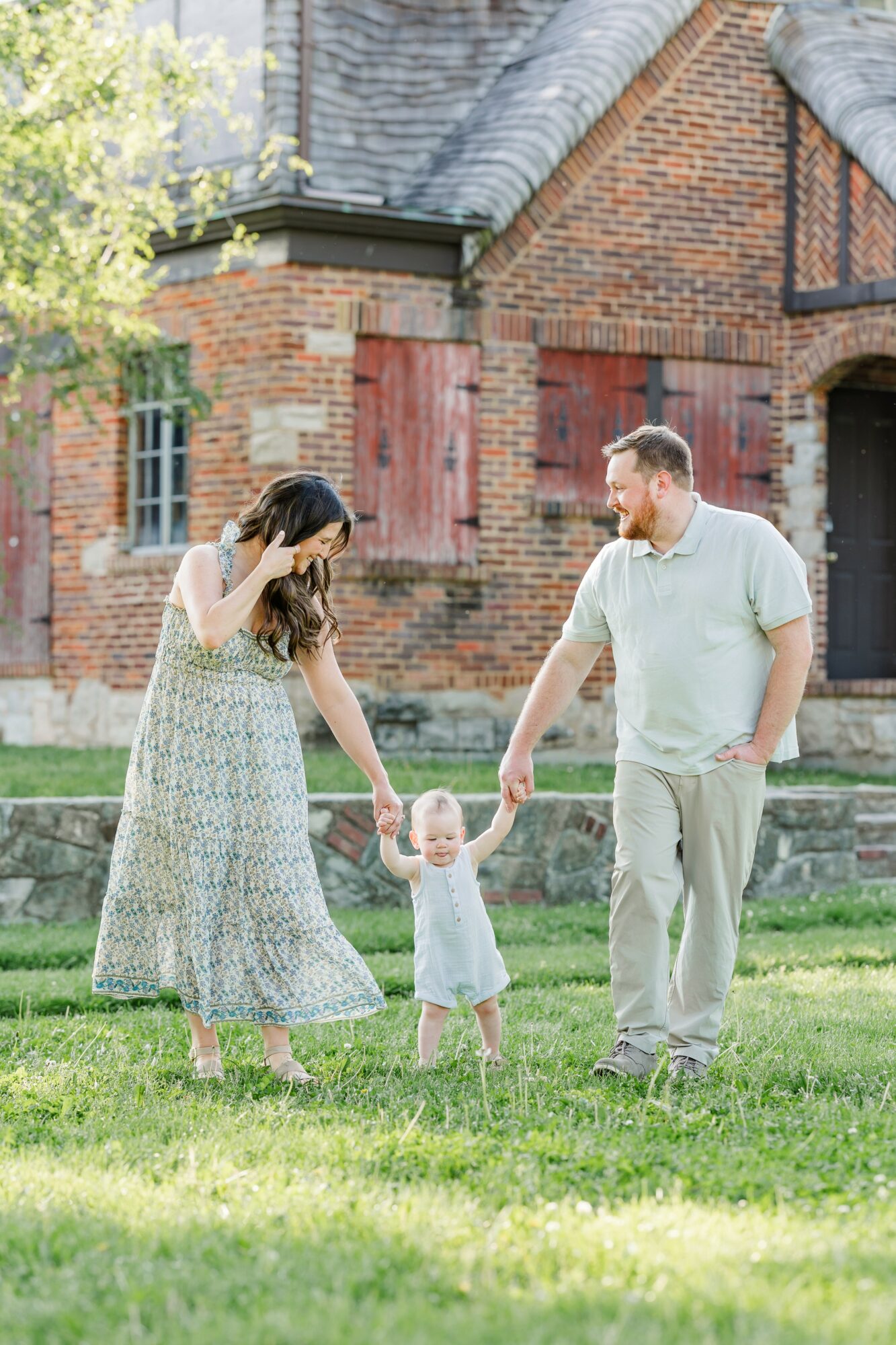 Family of three holding hands and walking on grass in front of a brick building, woman in a long dress, man in a white shirt, child in a white dress.