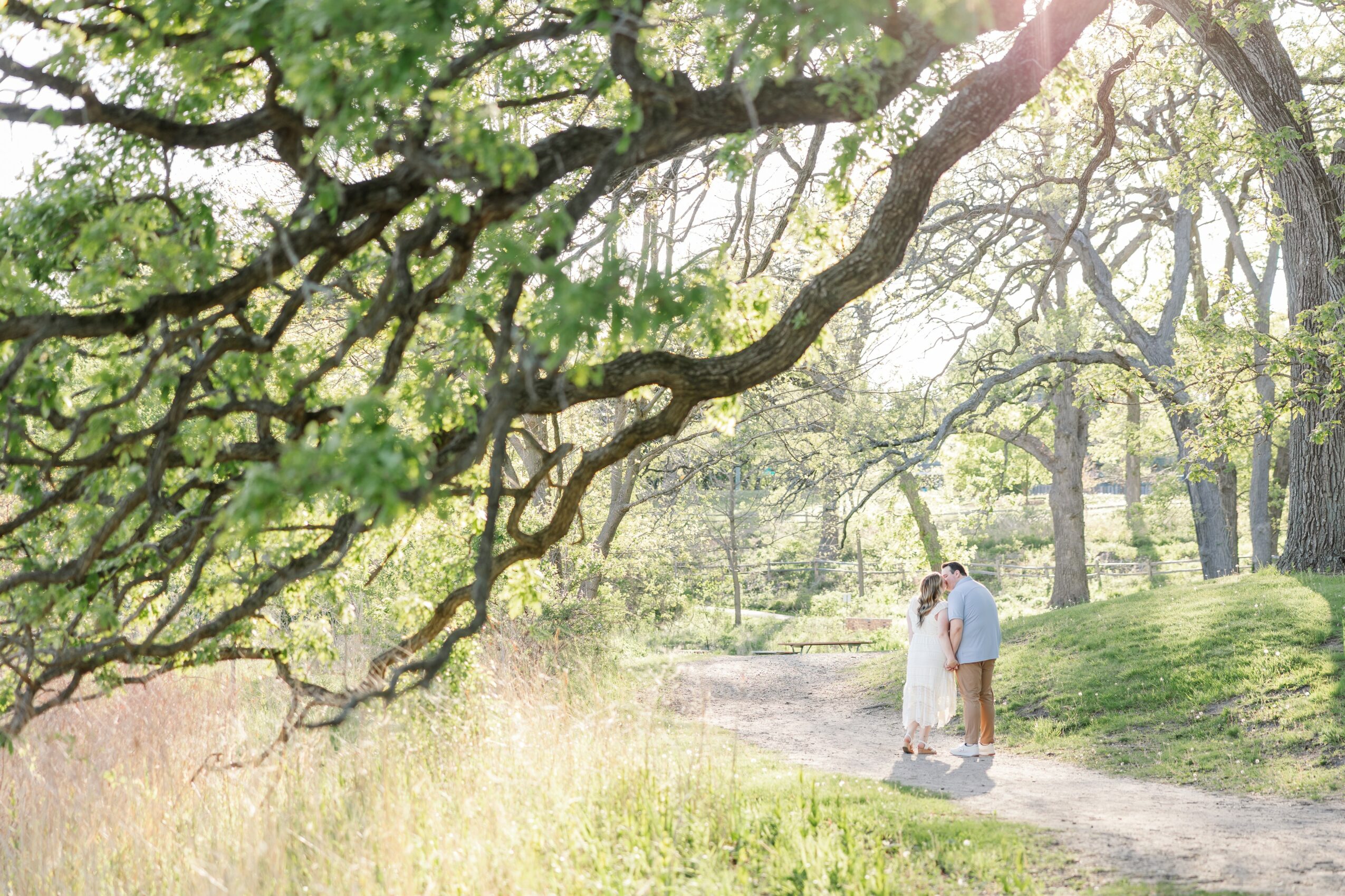 Two people walk along a dirt path in a park with trees and sunlight filtering through the branches.