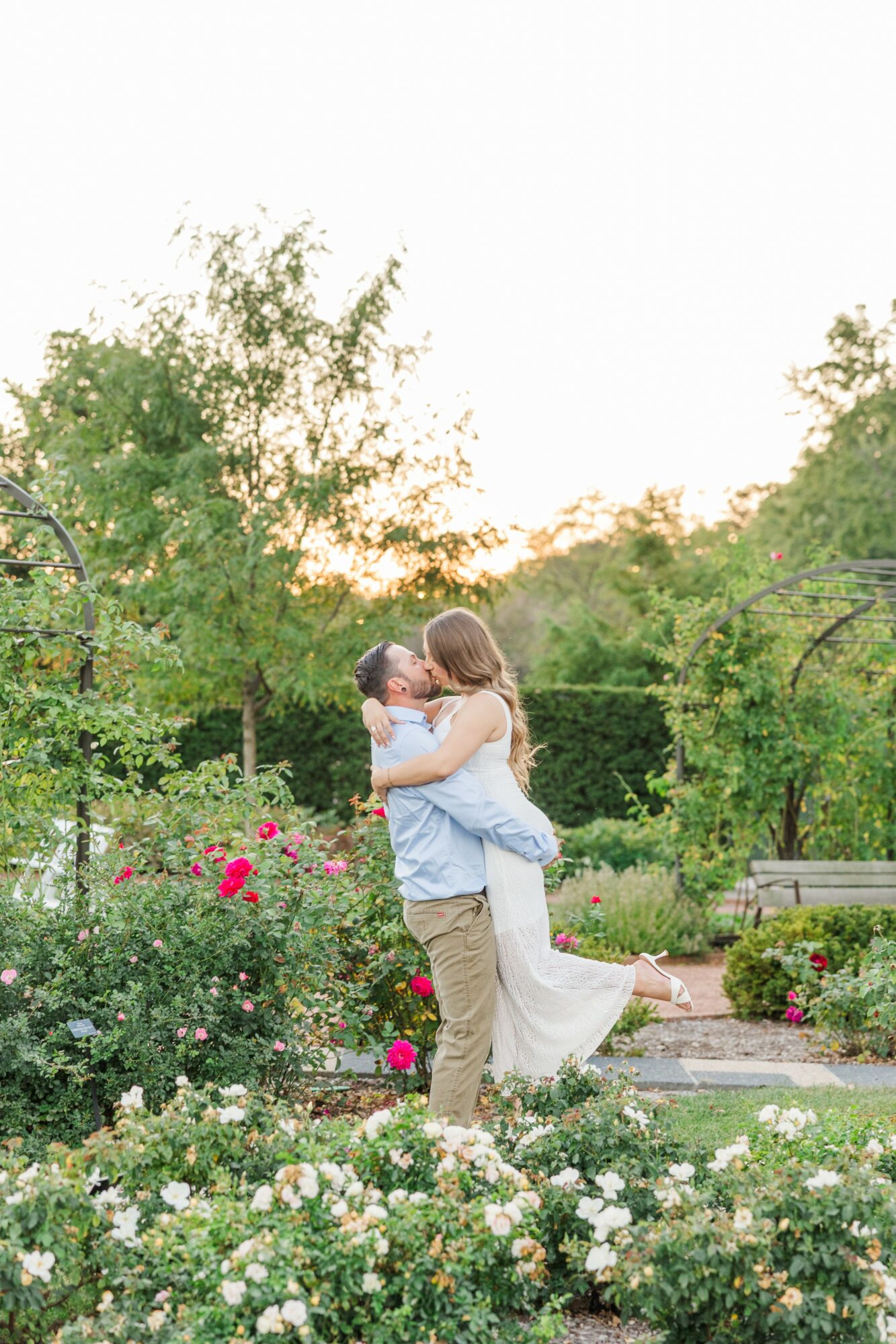 A couple embraces in a garden with flowers and trees during sunset, with a bench in the background.
