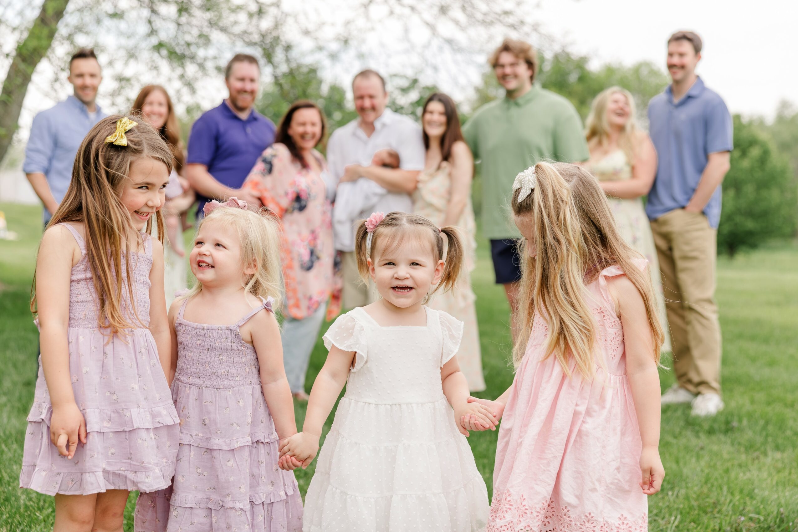 Group of children holding hands and smiling outdoors with adults in the background, trees, and grass.