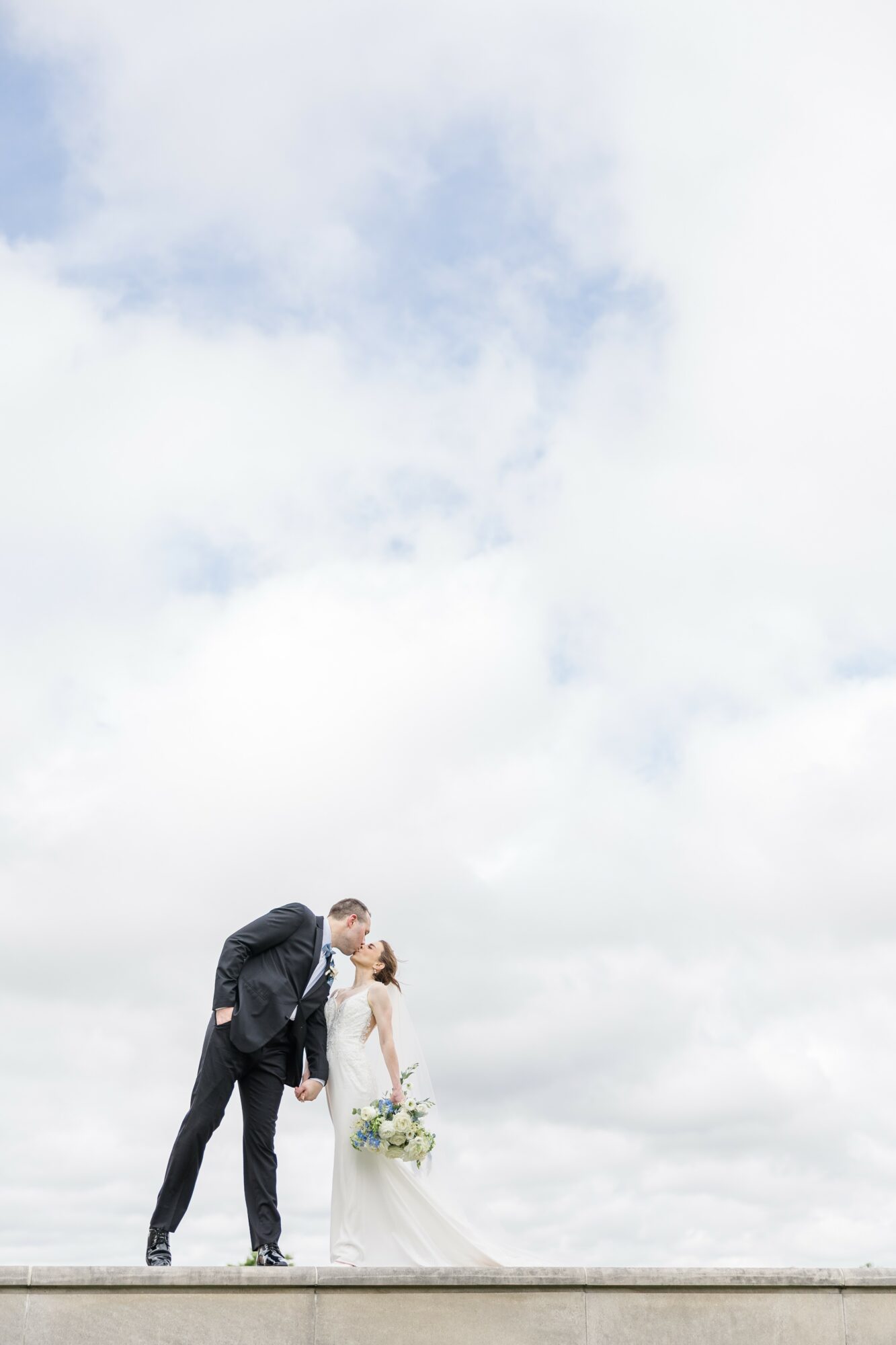 A bride and groom leaning toward each other, with the bride holding a bouquet, against a cloudy sky.