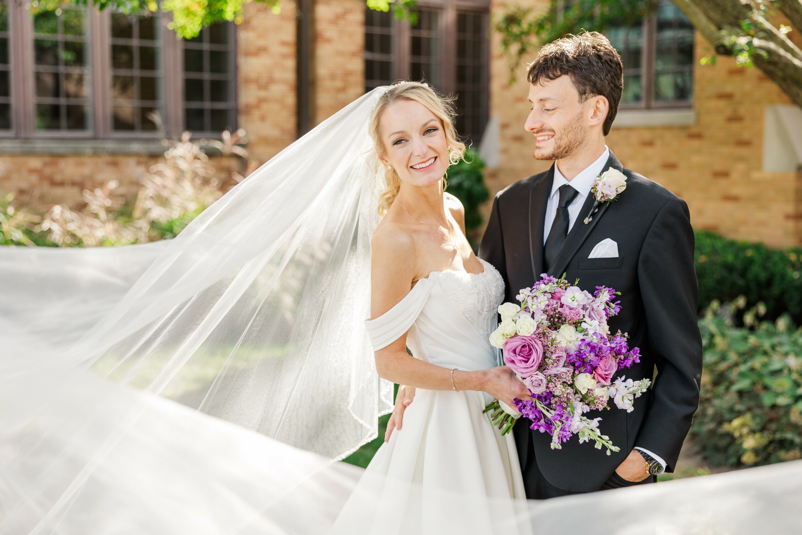 Bride and groom smiling outdoors, bride holding a bouquet of purple and white flowers, bride in white dress with veil, groom in dark suit