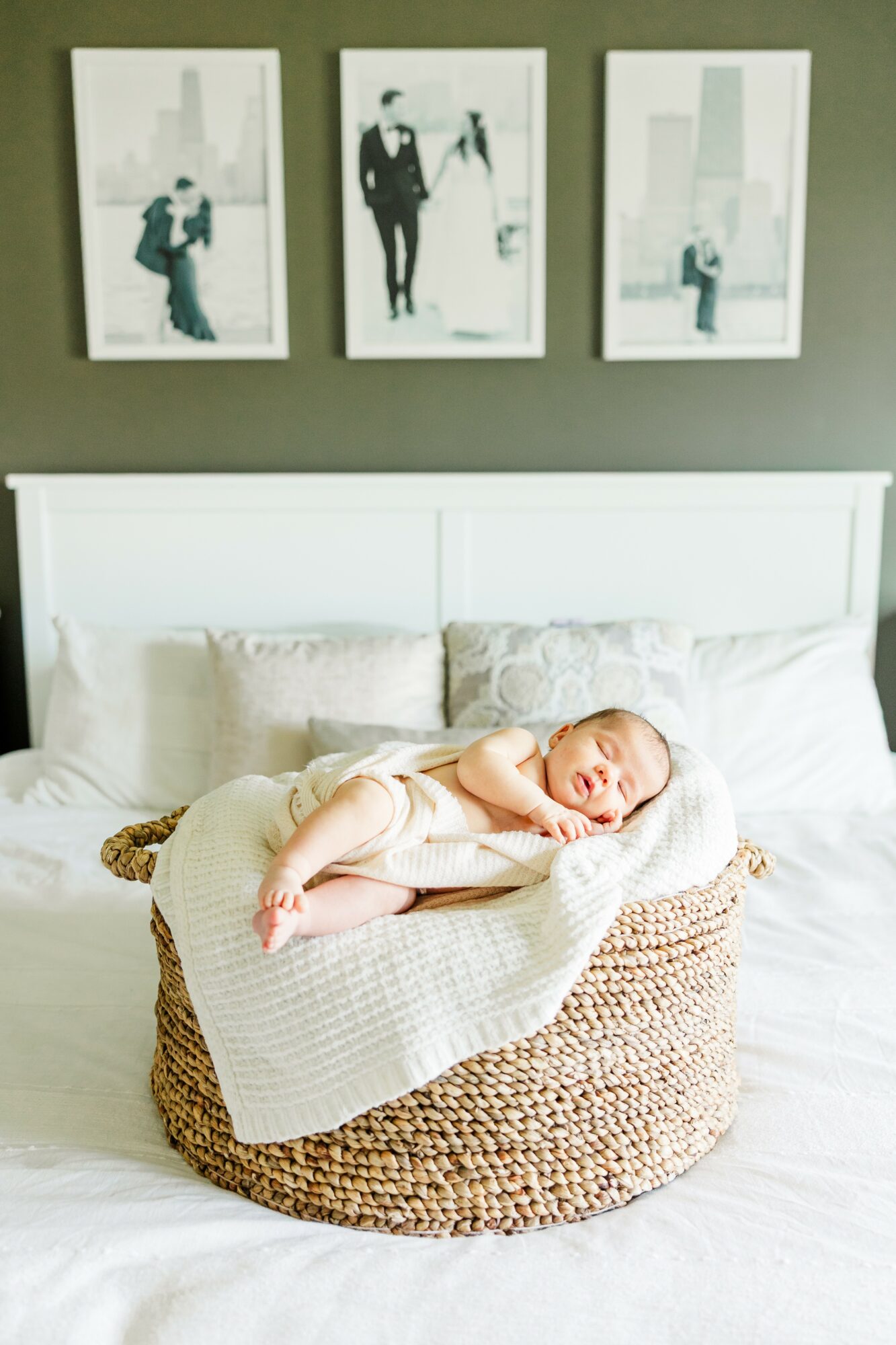 Baby sleeping on a blanket in a woven basket on a bed with pillows, above a white headboard, with three framed pictures on the wall.
