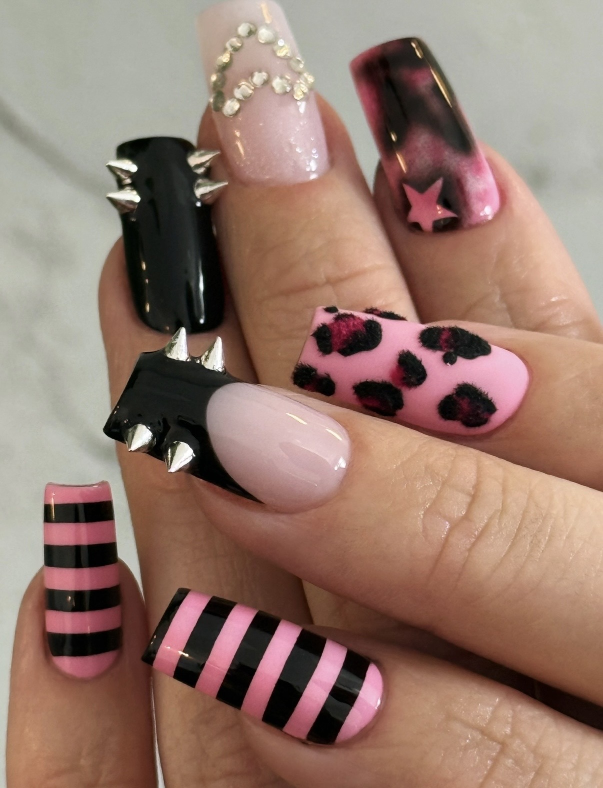 Close-up of a hand with decorated nails featuring pink, black, and animal print designs, some with spikes and stars.