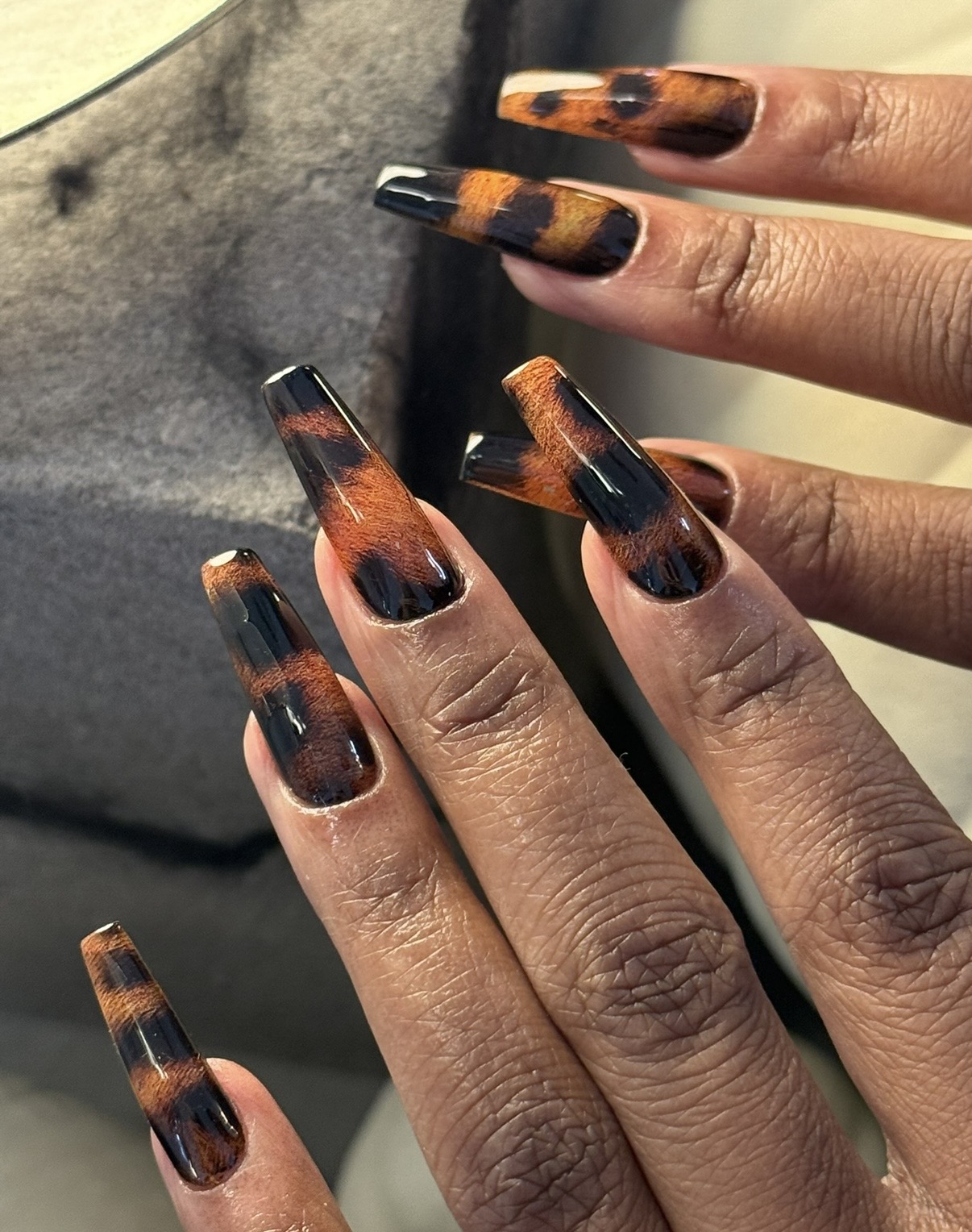 Close-up of hands with long, square-shaped nails painted in a tortoiseshell pattern, against a dark background.