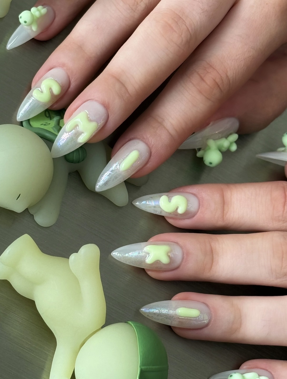 Hands with long, almond-shaped nails painted with a light, iridescent base and green, white, and yellow decorative accents, surrounded by small green and white decorative objects.