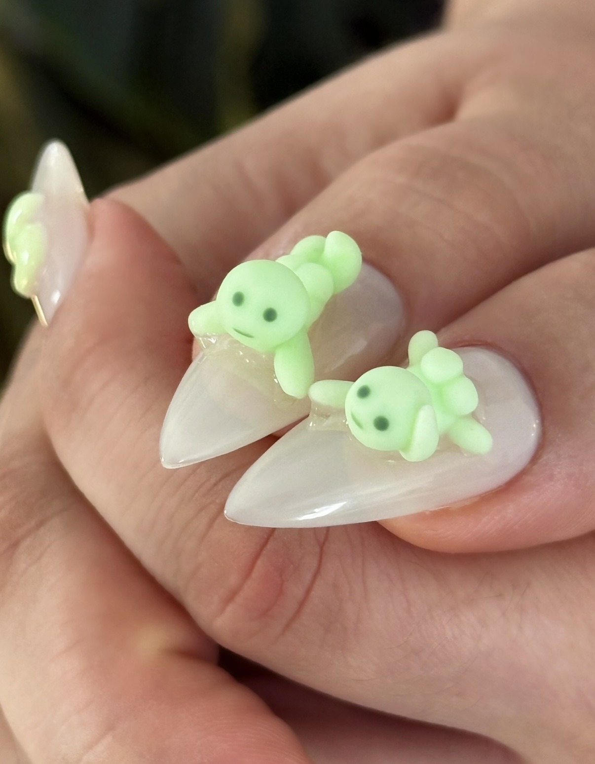 Close-up of a hand with decorated nails and small green turtle-shaped nail art accessories, with a blurred background.
