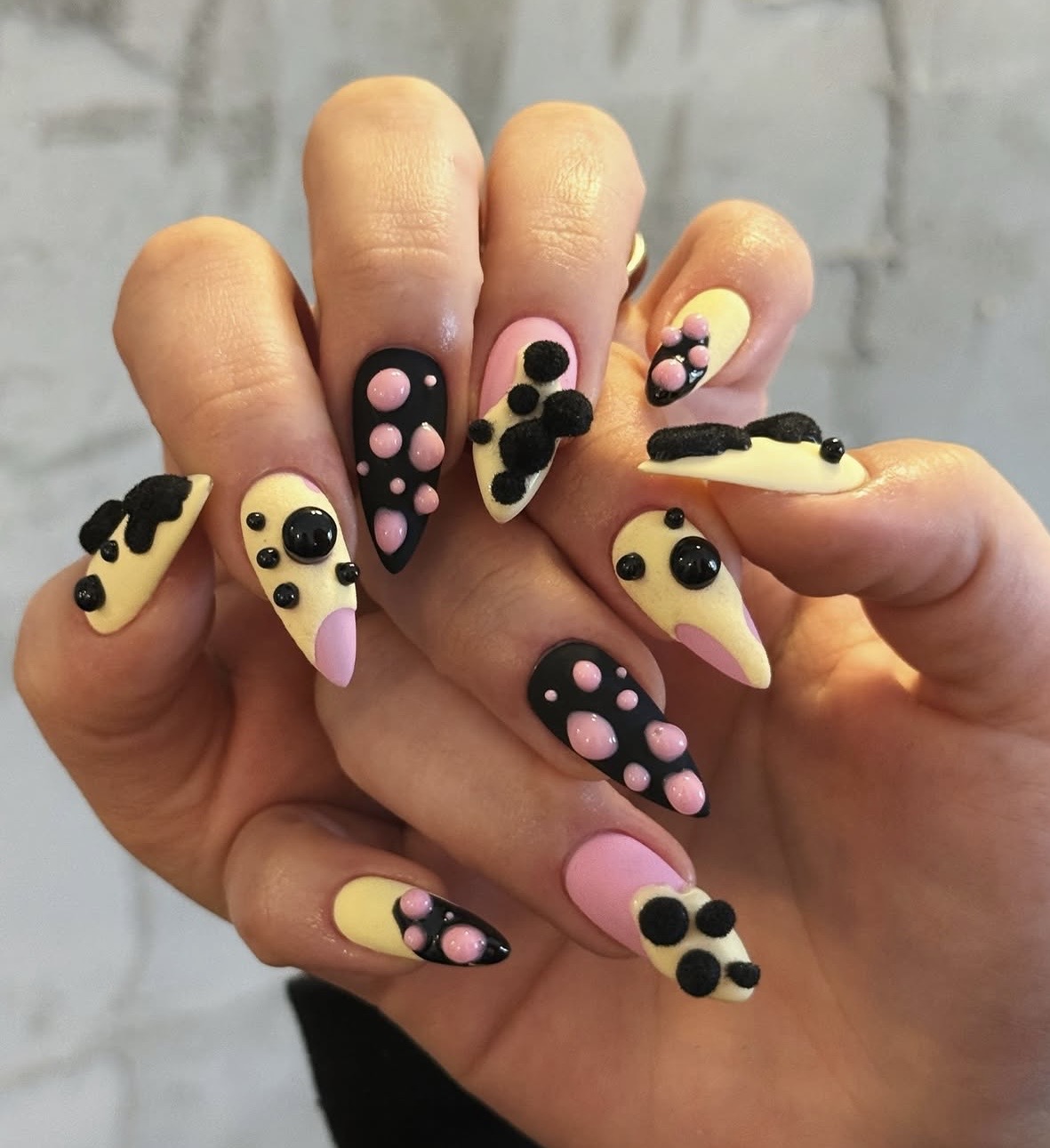 Hand with nails painted in pastel yellow, pink, and black, decorated with polka dots and bow designs, against a gray background.