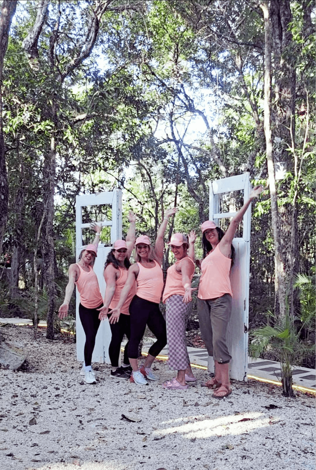 Group of six women in pink shirts and hats standing outdoors among trees, posing near white rectangular frames.