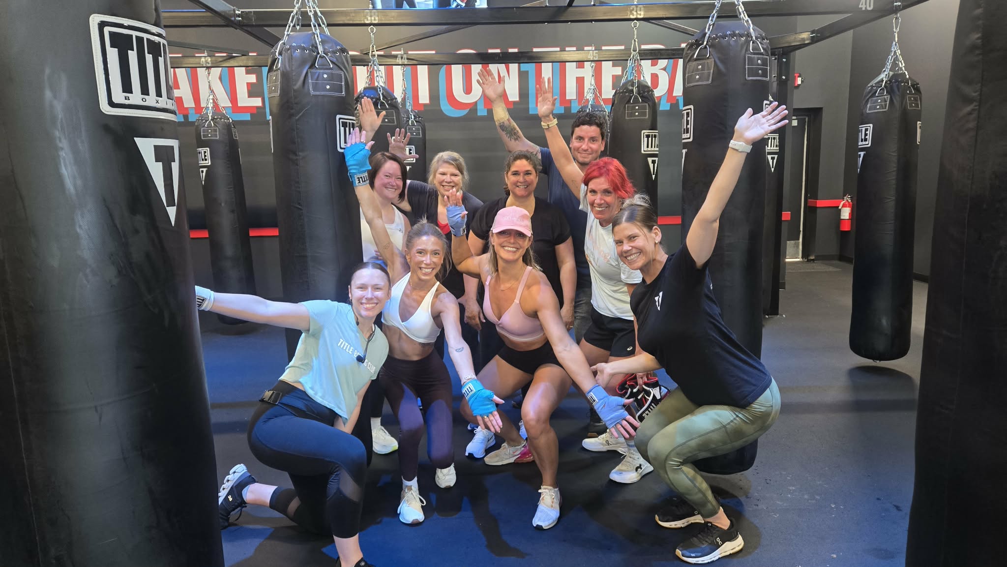 Group of people in a boxing gym, smiling and posing with boxing gloves and equipment.