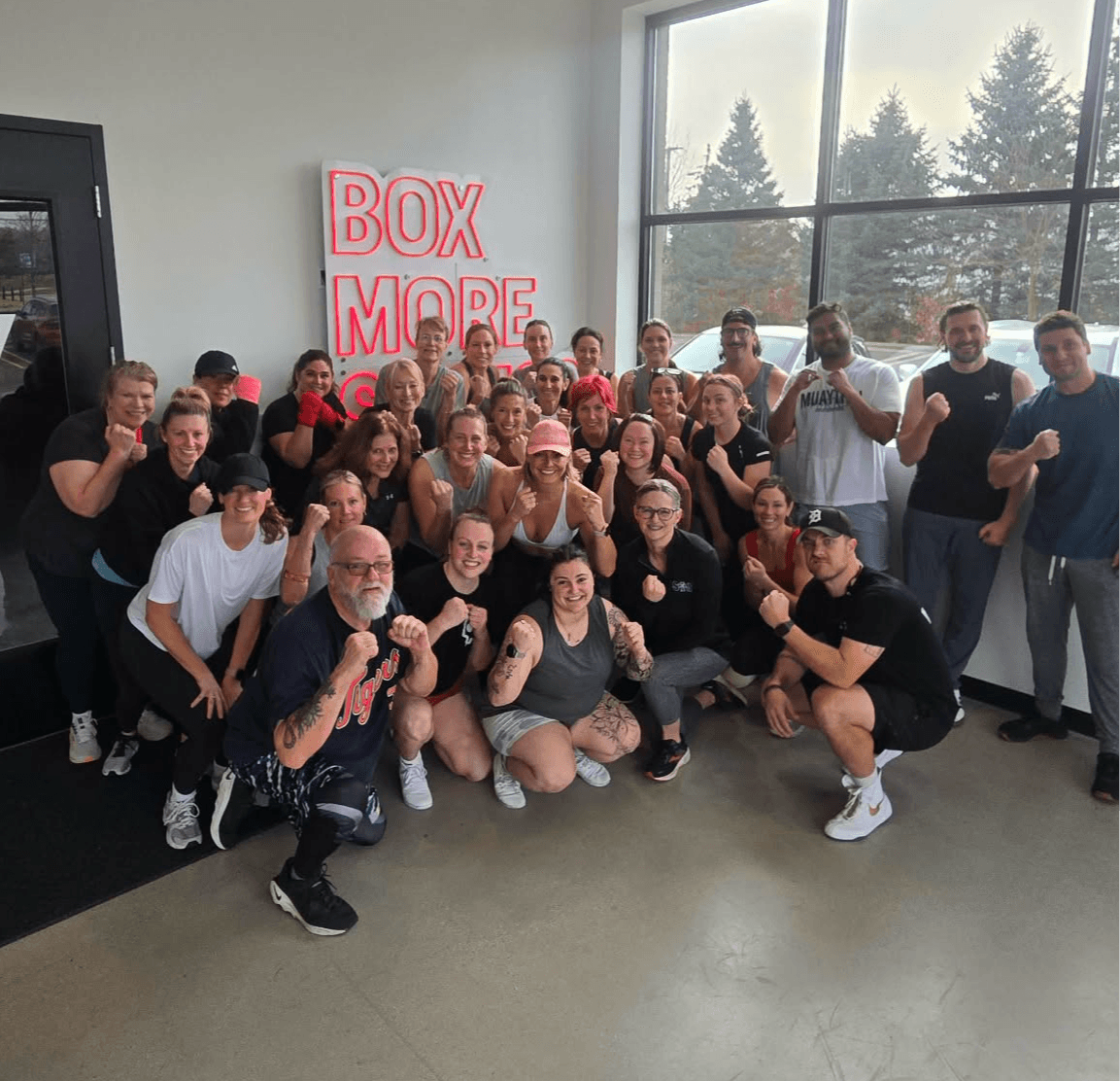 Group of people posing indoors with a window and a sign that says 'BOX MORE' in the background.