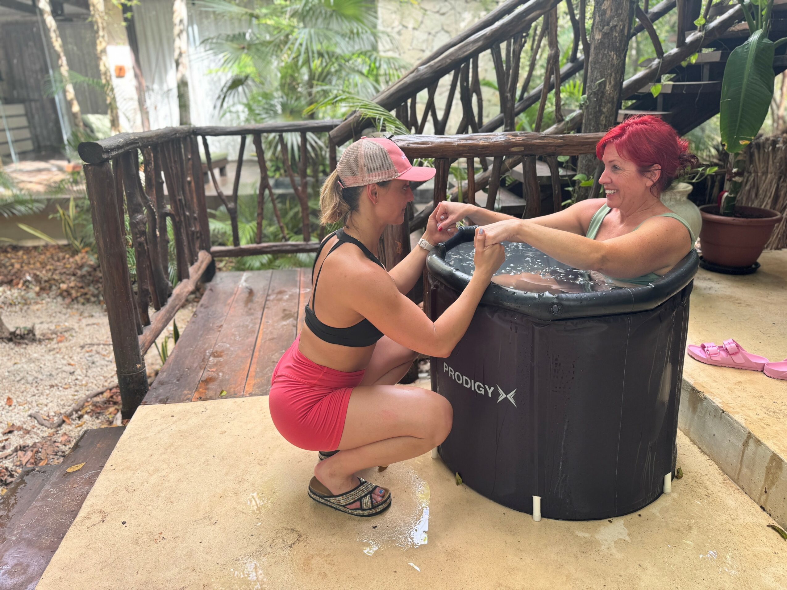 Two women in a hot tub outdoors, one with red hair and the other with a cap, holding hands and smiling.