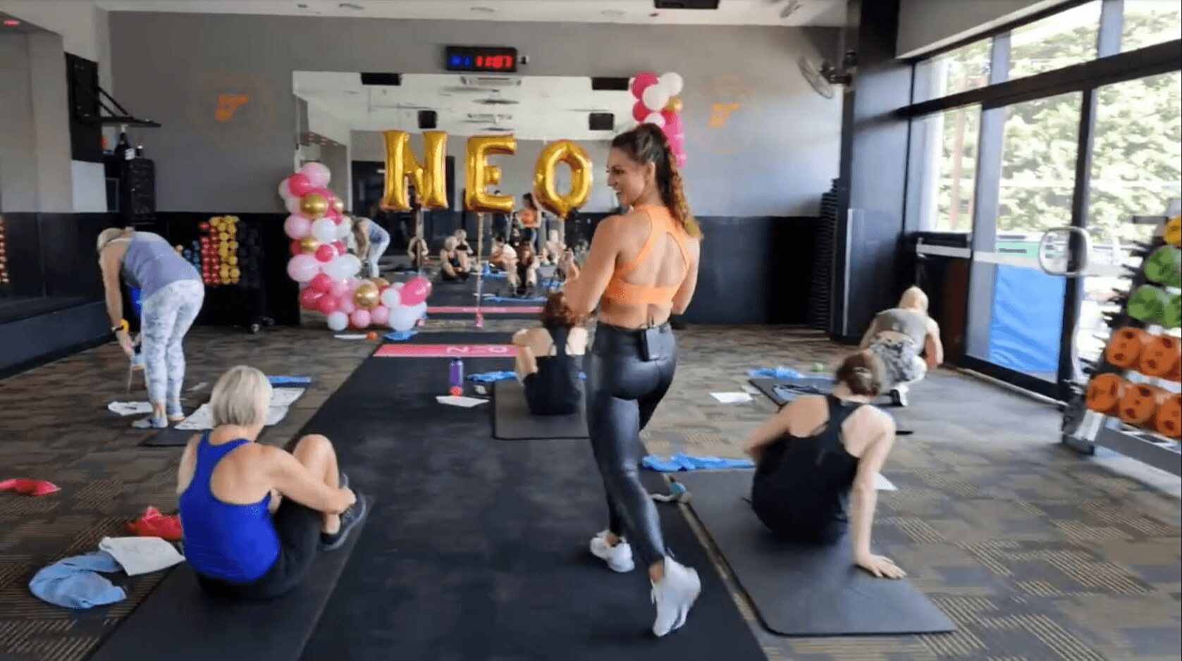 Group of women and children in a fitness studio with balloons and decorations, some sitting and others standing, engaging in activities.
