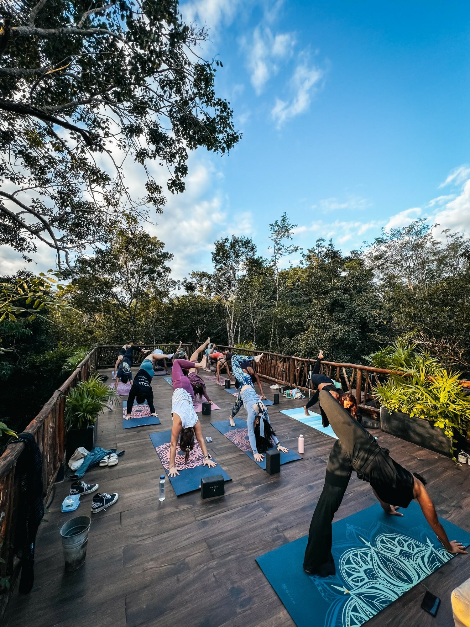 People practicing yoga outdoors on a wooden deck with trees and blue sky in the background.