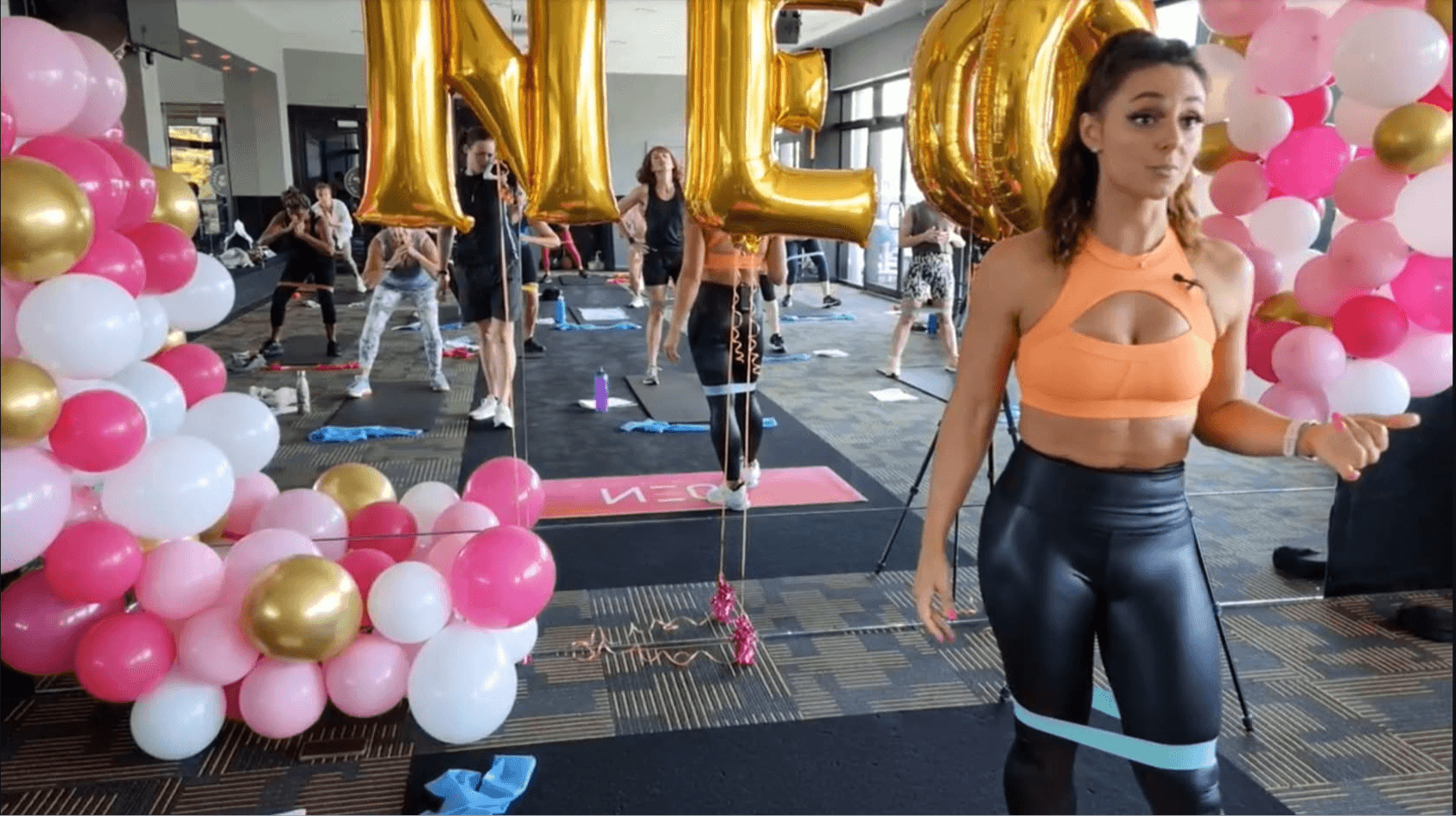 Indoor fitness class with balloons, large gold balloons spelling 'NEW', and women exercising with resistance bands.