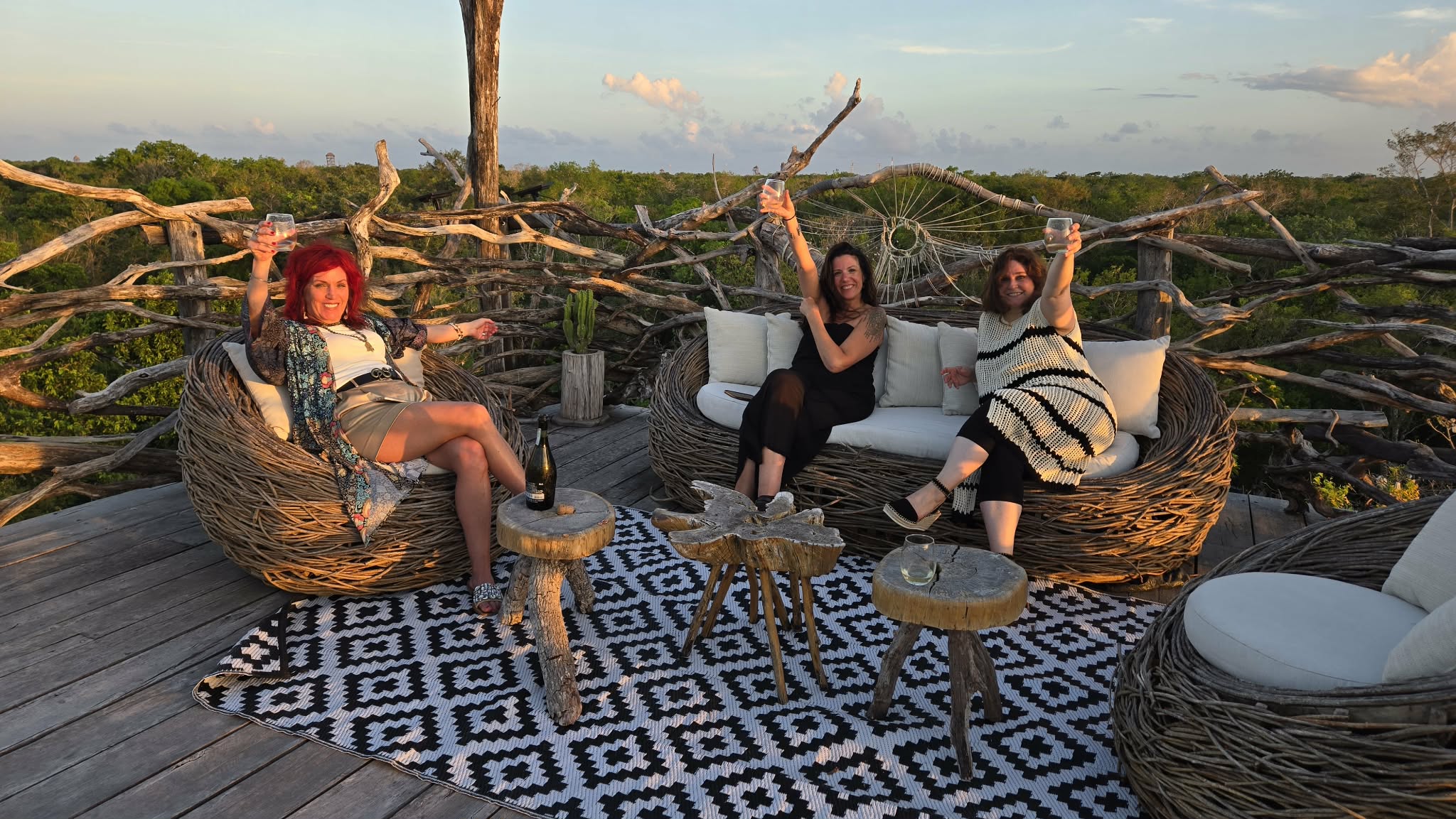 Three women sit on a wicker sofa and chairs outdoors on a wooden deck with a patterned rug, holding sticks, with a landscape background.