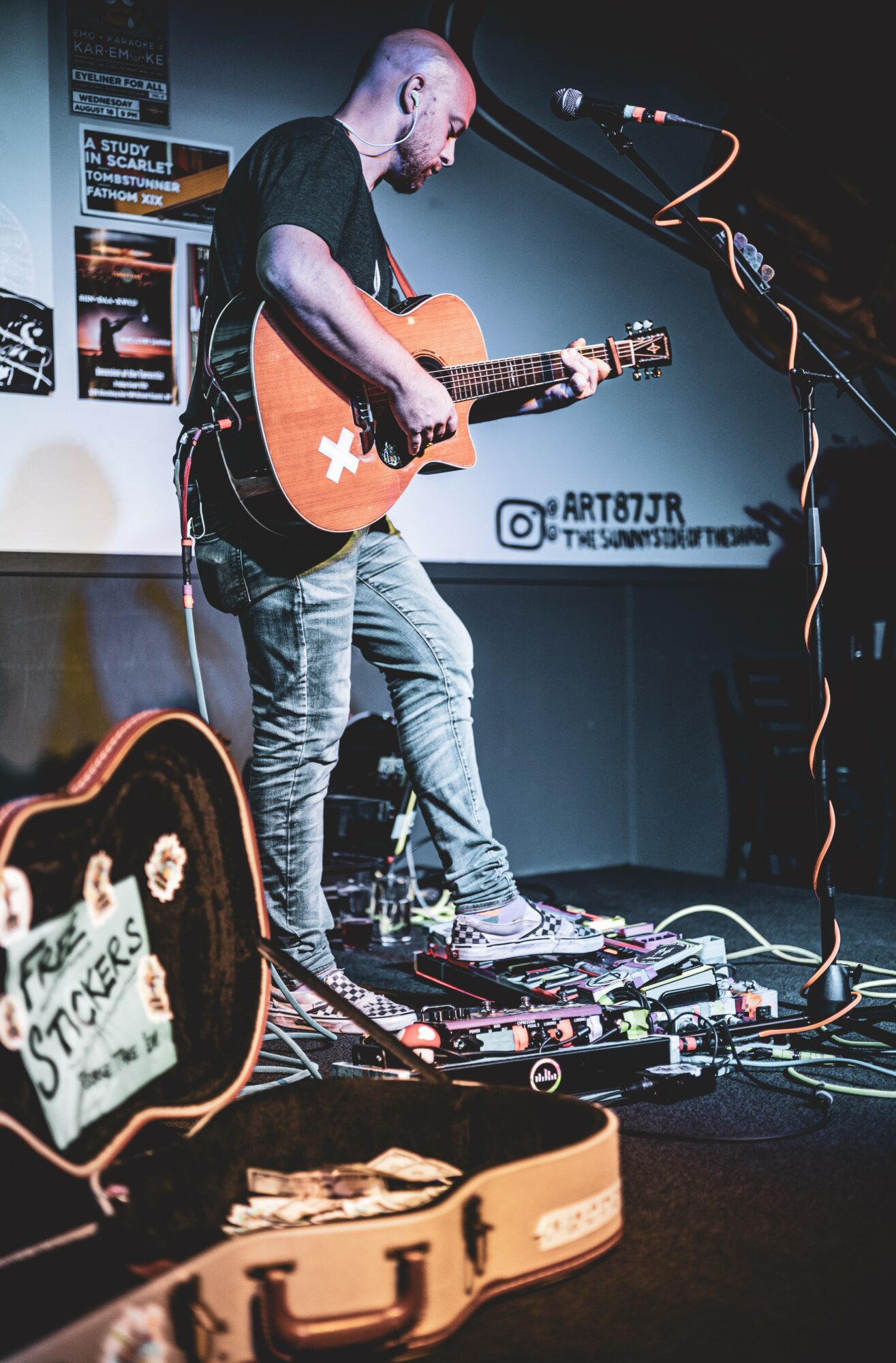 Musician playing guitar on stage with equipment and a sign in the background, performing in a dimly lit venue.