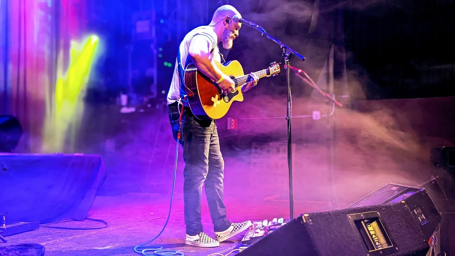 Person playing guitar on stage with colorful lights and fog effects, microphone stand nearby.