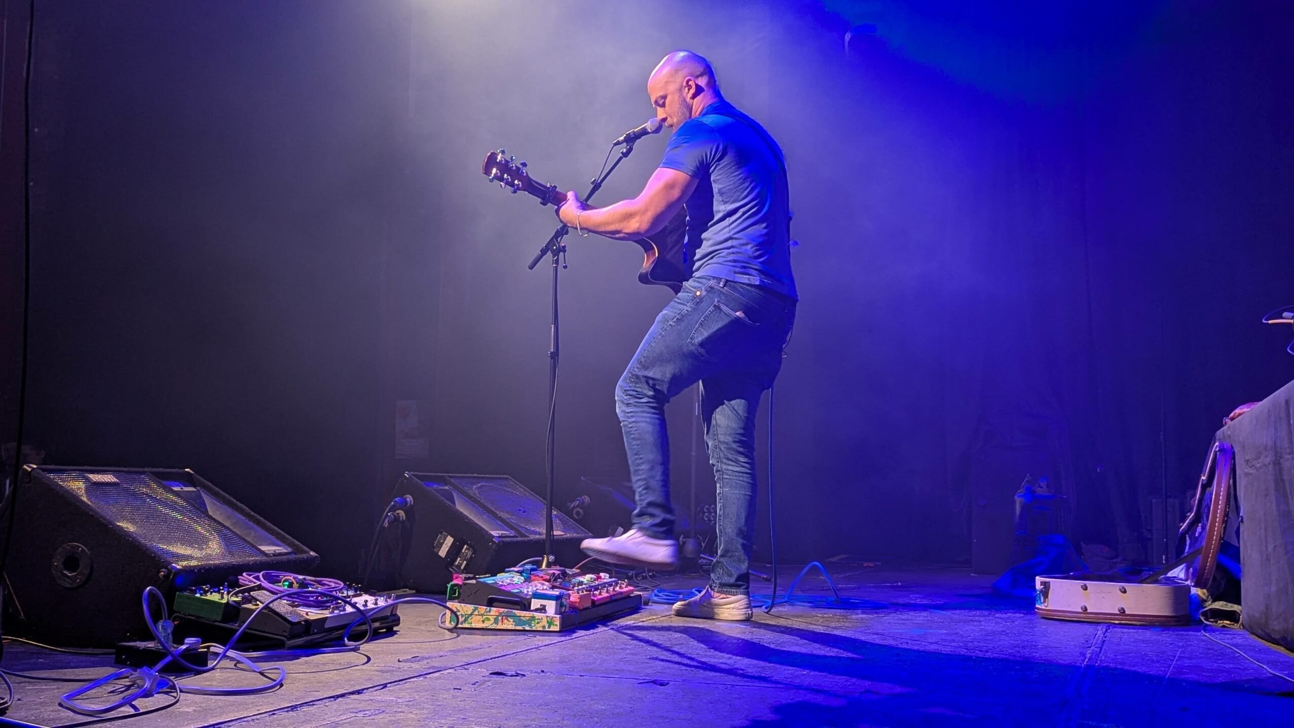 Person playing guitar on stage with equipment and blue lighting, facing right, mid-performance.