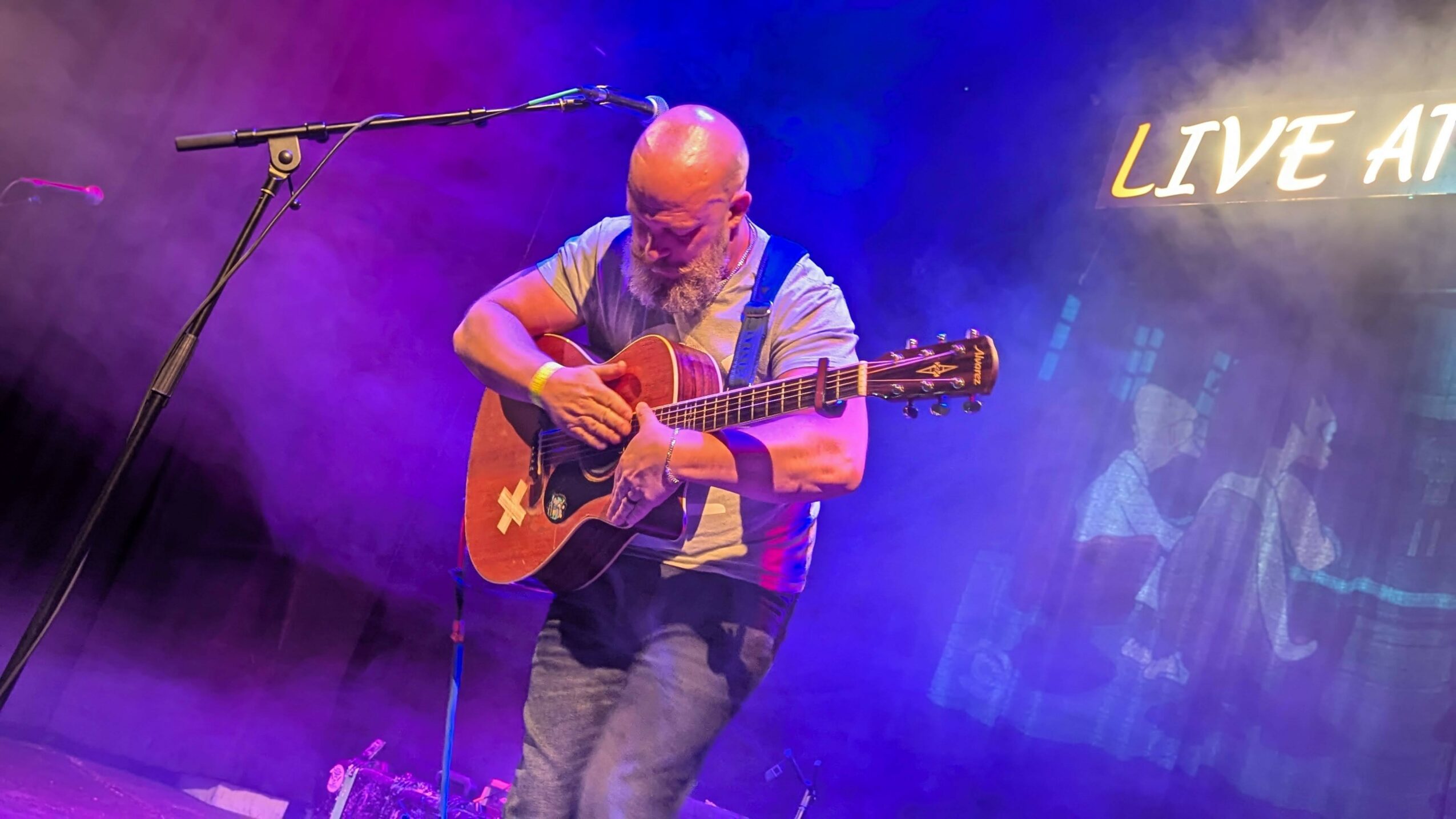 Bald man playing guitar on stage with purple and blue lighting, and a partially visible sign reading 'LIVE AT'.