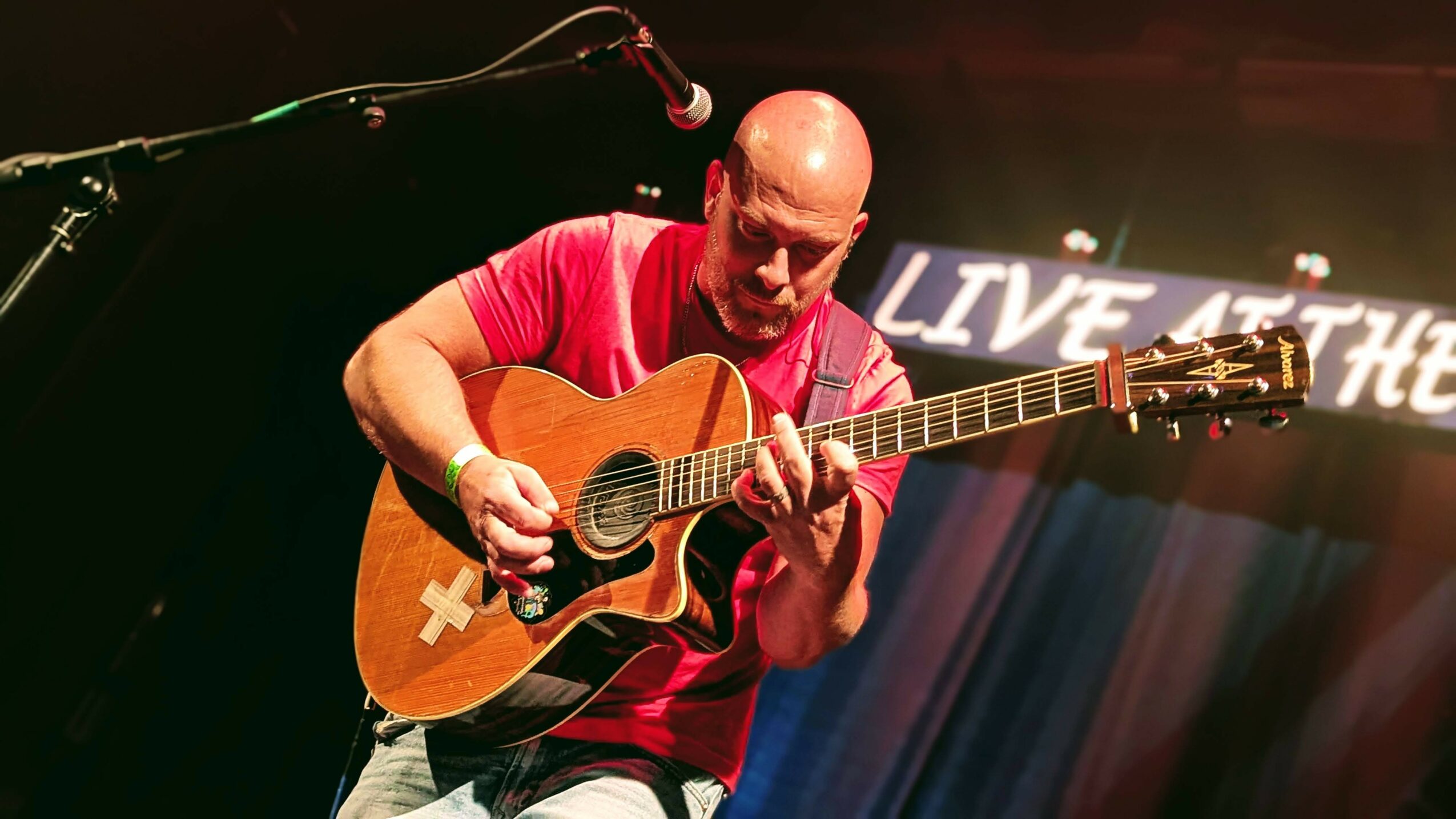 Bald man playing an acoustic guitar on stage with a microphone, stage lights, and a sign in the background.
