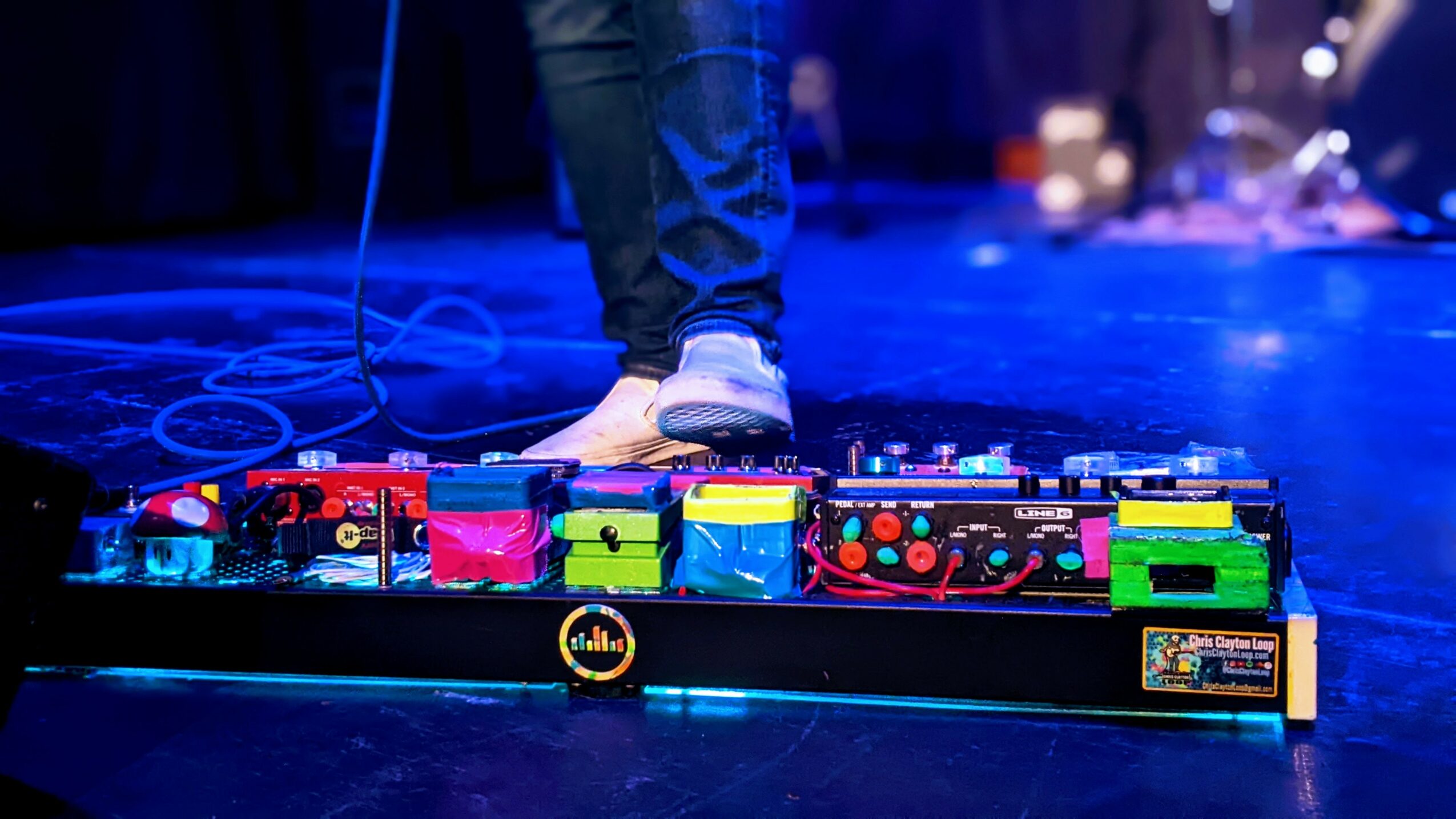 Person standing on stage with colorful guitar effects pedals, wearing jeans and sneakers, stage lights in background.
