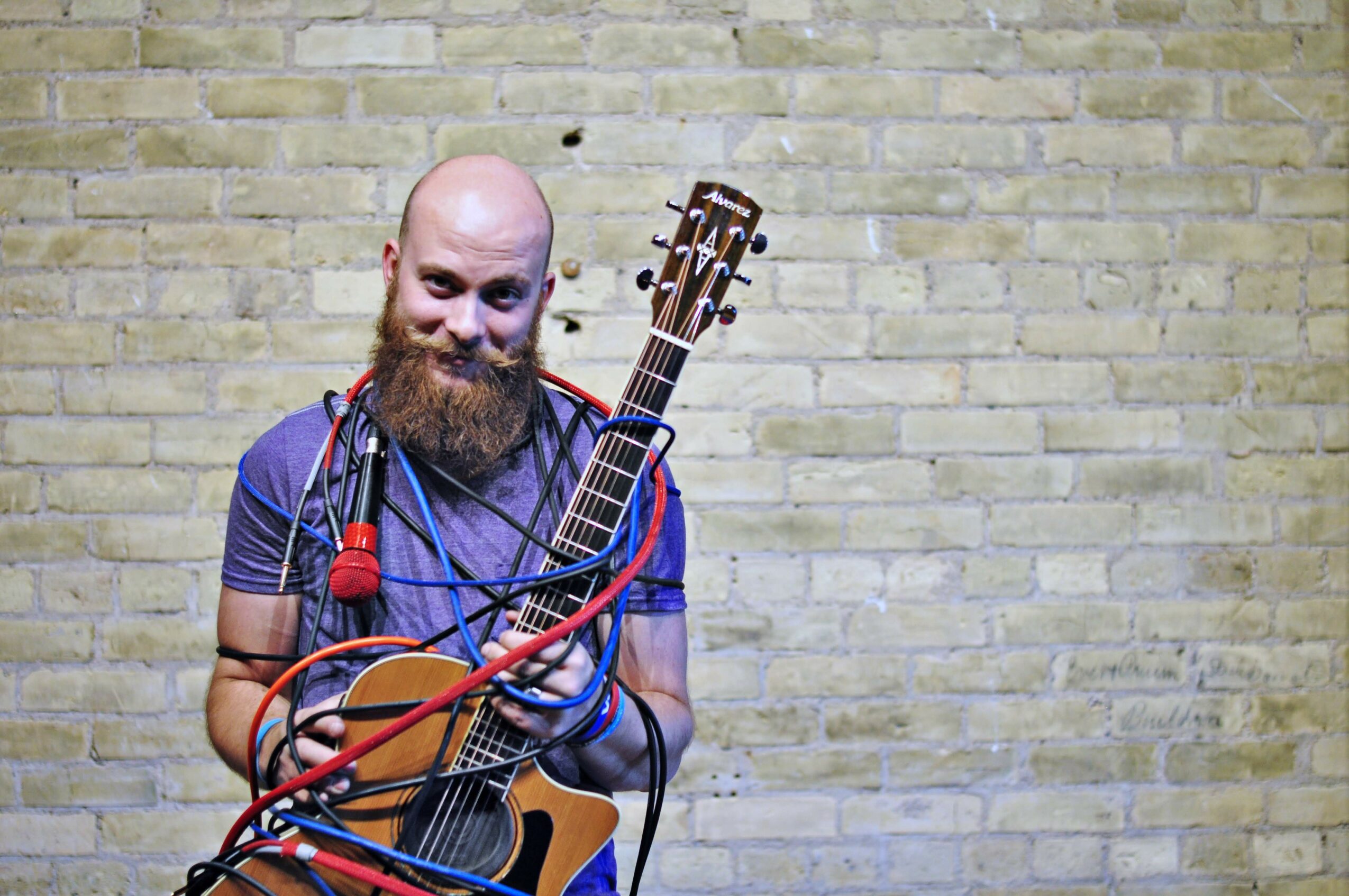 Bearded man holding an acoustic guitar with cables, standing against a brick wall, smiling.