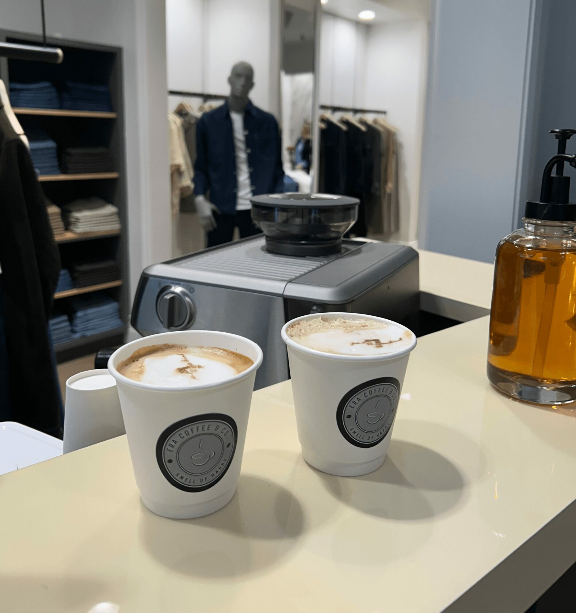 Two cups of coffee on a counter with a soap dispenser and a machine in the background, clothing store interior.
