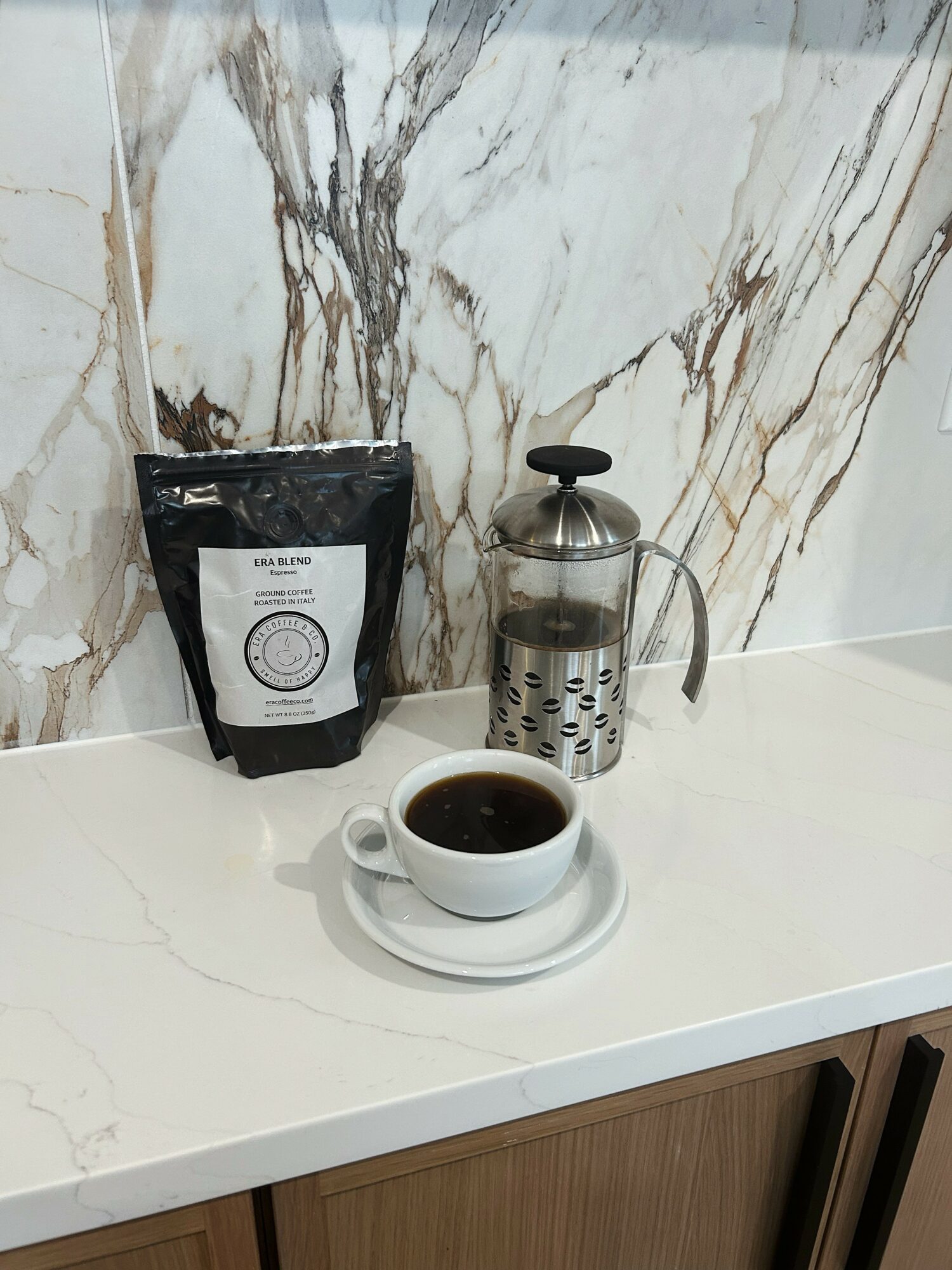 Cup of coffee on a saucer, French press, and coffee bag on a white countertop with marble wall background.